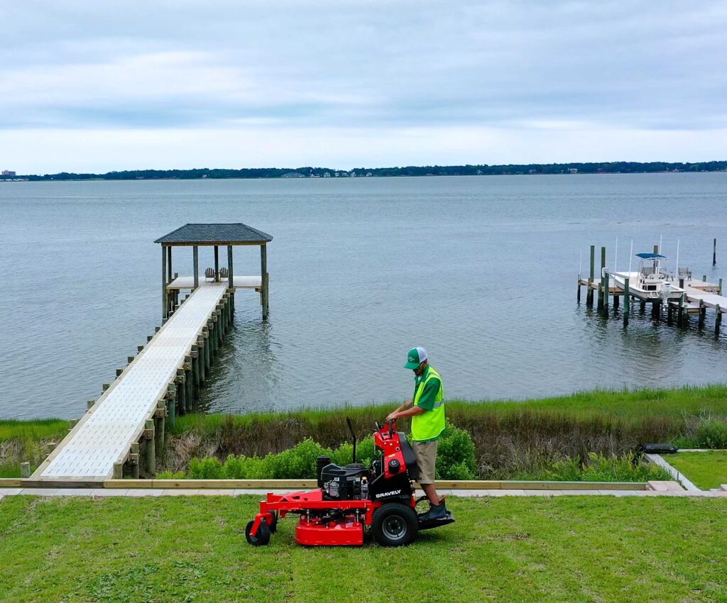 A person in a high-visibility vest operates a lawnmower near a dock by a calm water body under a cloudy sky.