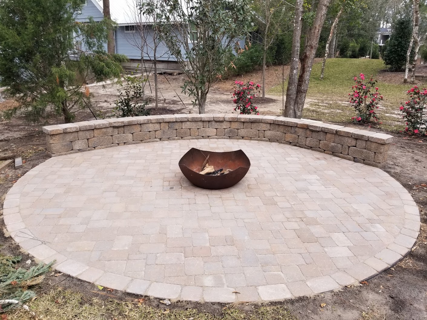 A circular patio with a metal fire pit, surrounded by a stone wall and trees, features fresh plants and a house in the background.