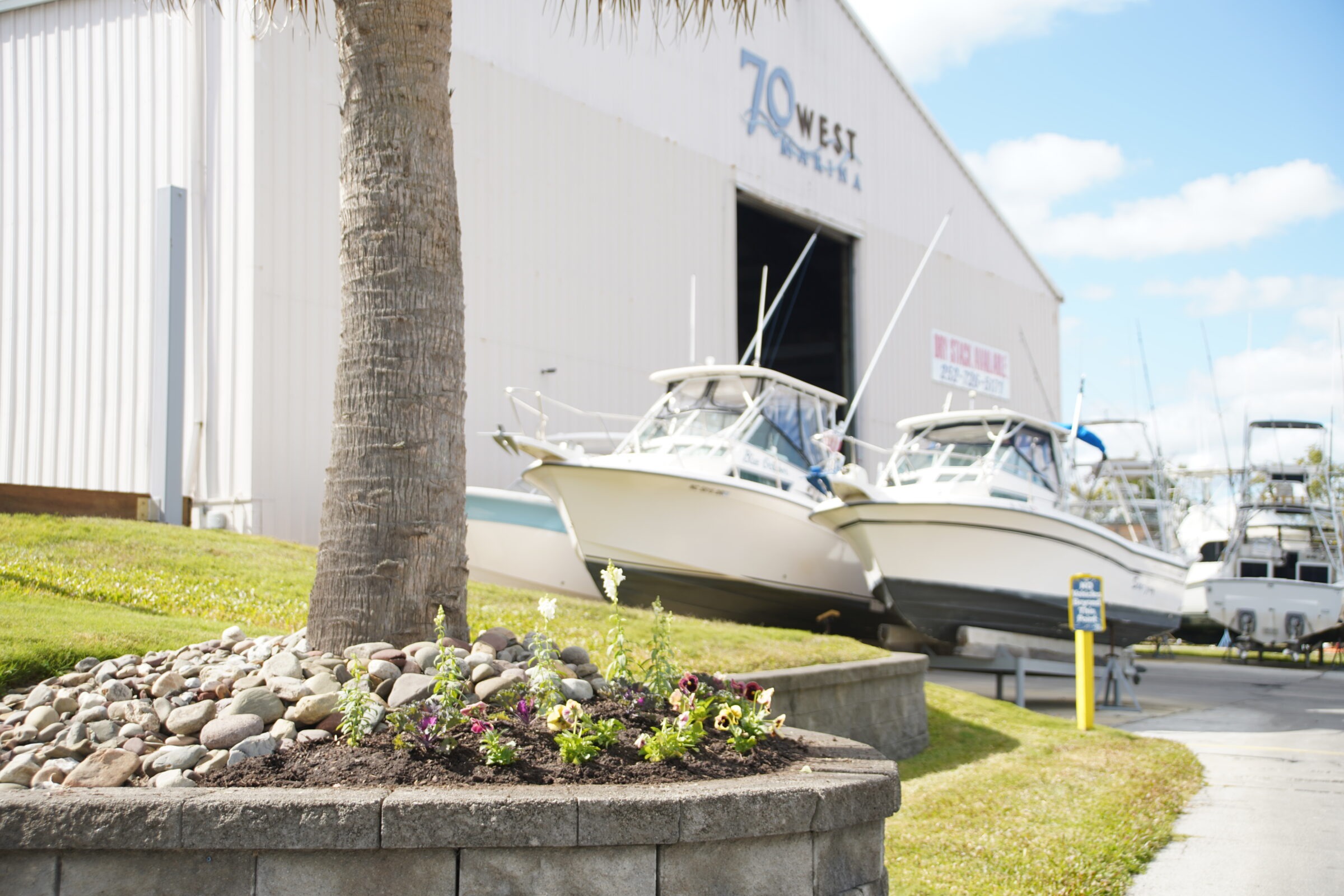 Boats are stored outside a large warehouse at 70 West Marina, with a palm tree and flower bed in the foreground.
