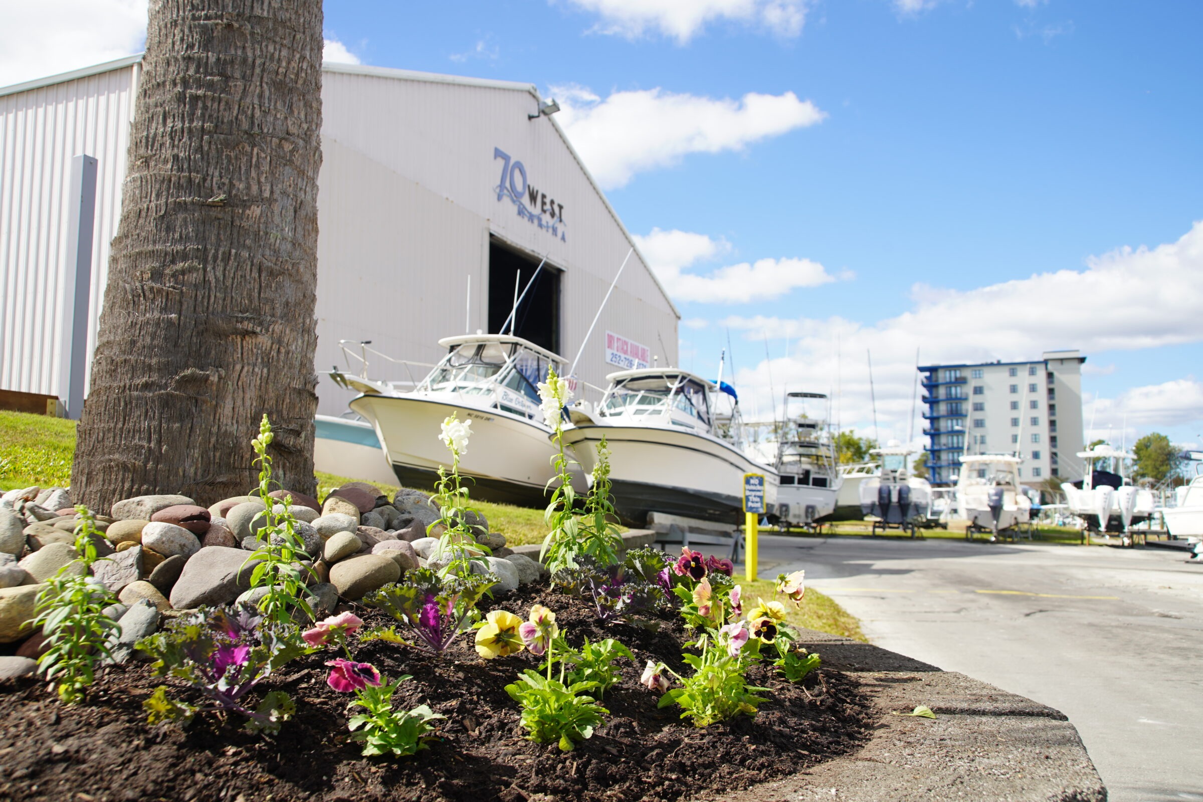 Boats on trailers are parked outside a large warehouse labeled "70 West Marina." Flowers and rocks decorate the foreground under a blue sky.