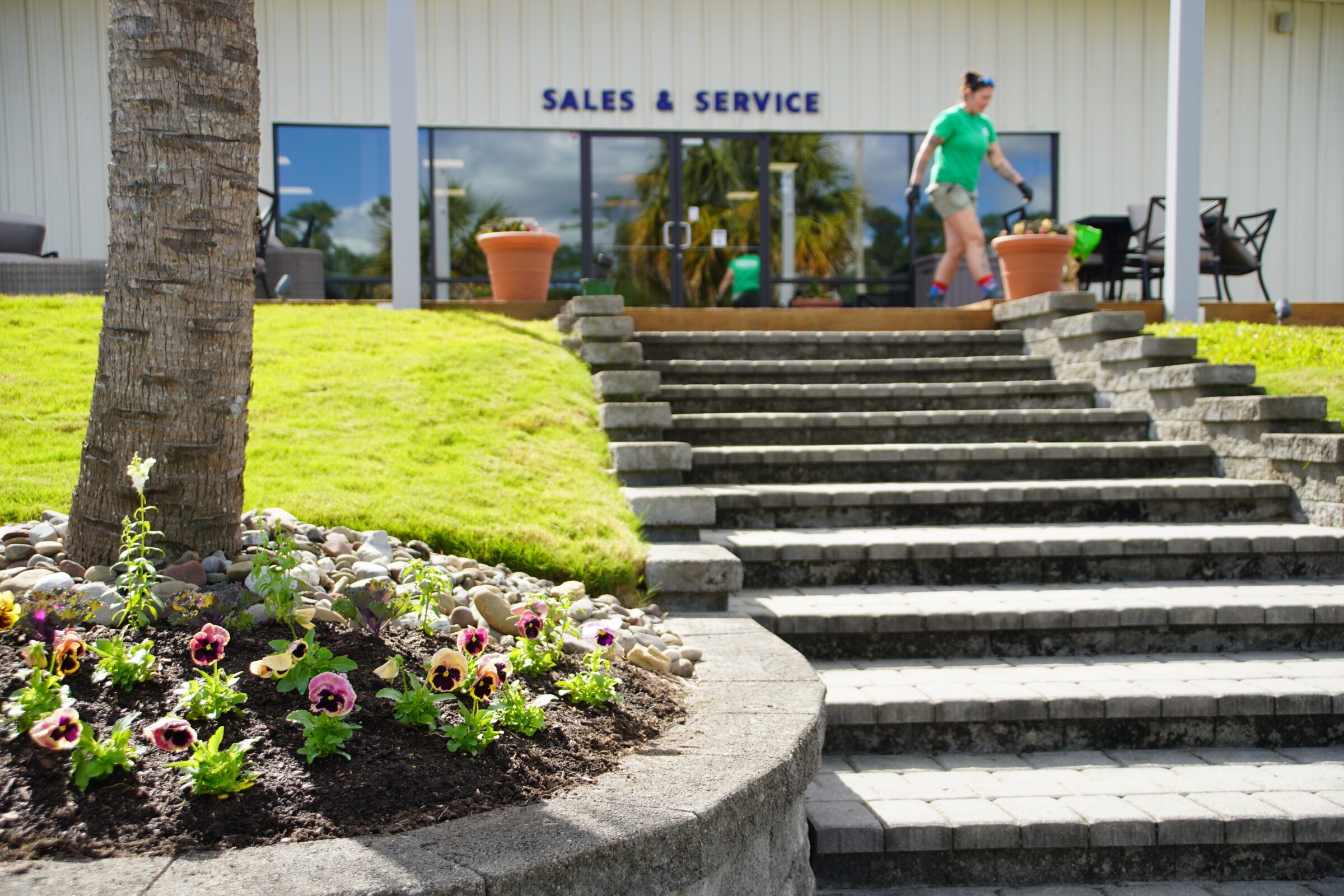 A person walks past a "Sales & Service" building entrance, with stone steps and vibrant flower bed in the foreground.