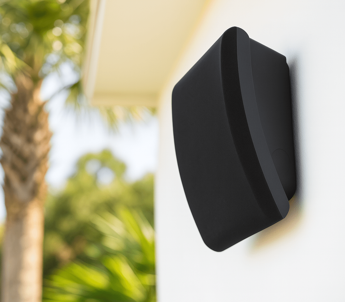 A black outdoor speaker mounted on a white wall, surrounded by blurred palm trees, under a clear sky. Modern and minimalist design.