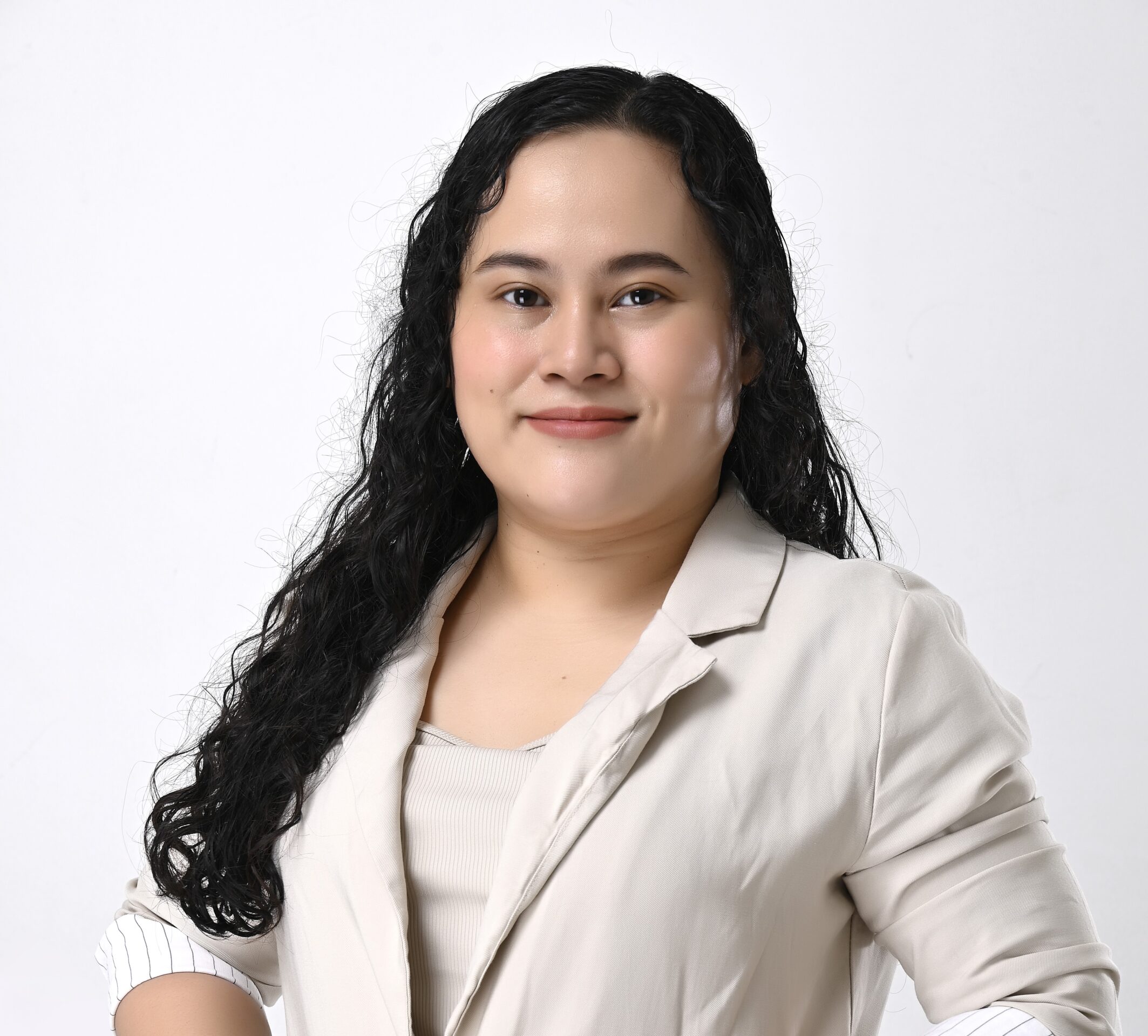A person with long, curly hair wears a light blazer, smiling against a plain white background. No landmarks or historical buildings visible.
