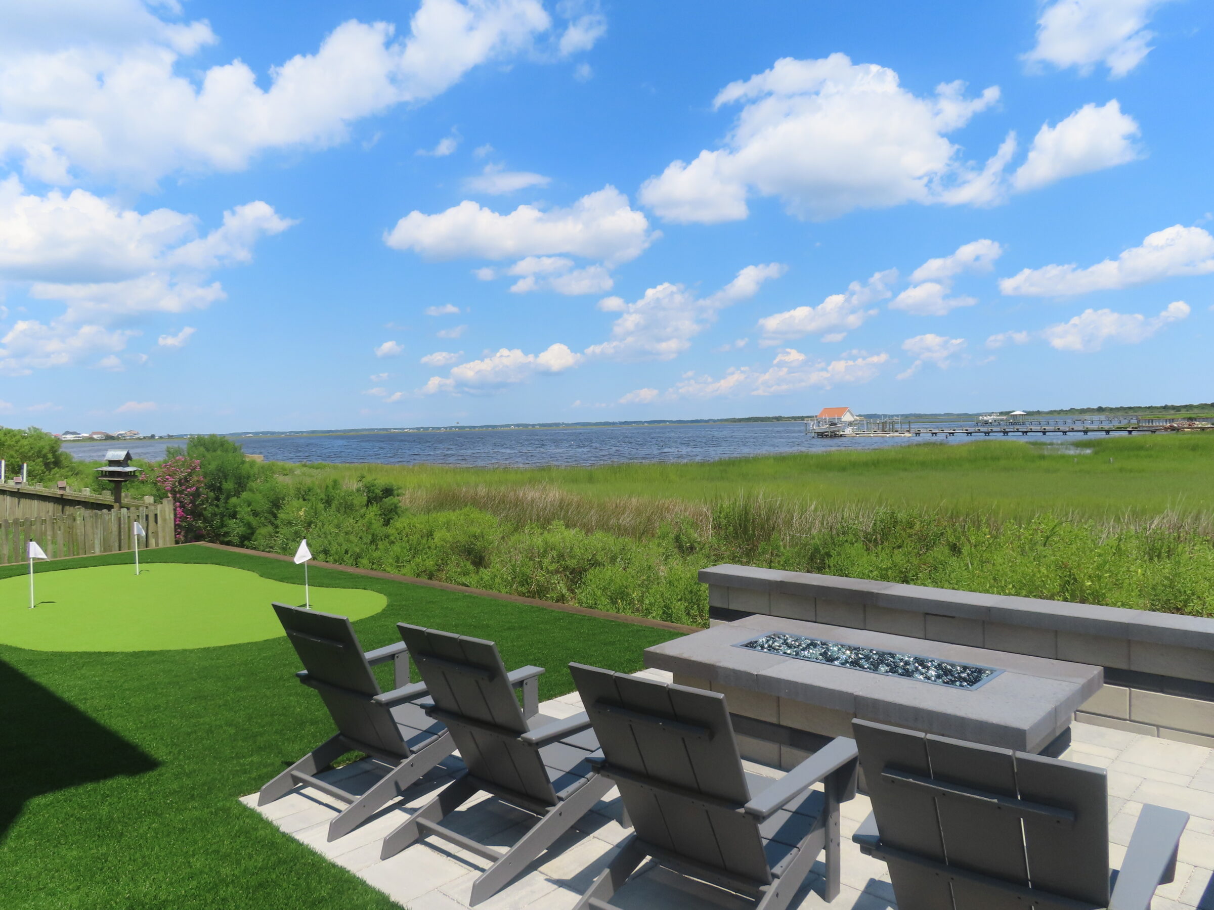 Backyard with golf putting green, lounge chairs, fire pit, and water view. Blue sky with clouds and distant buildings visible.