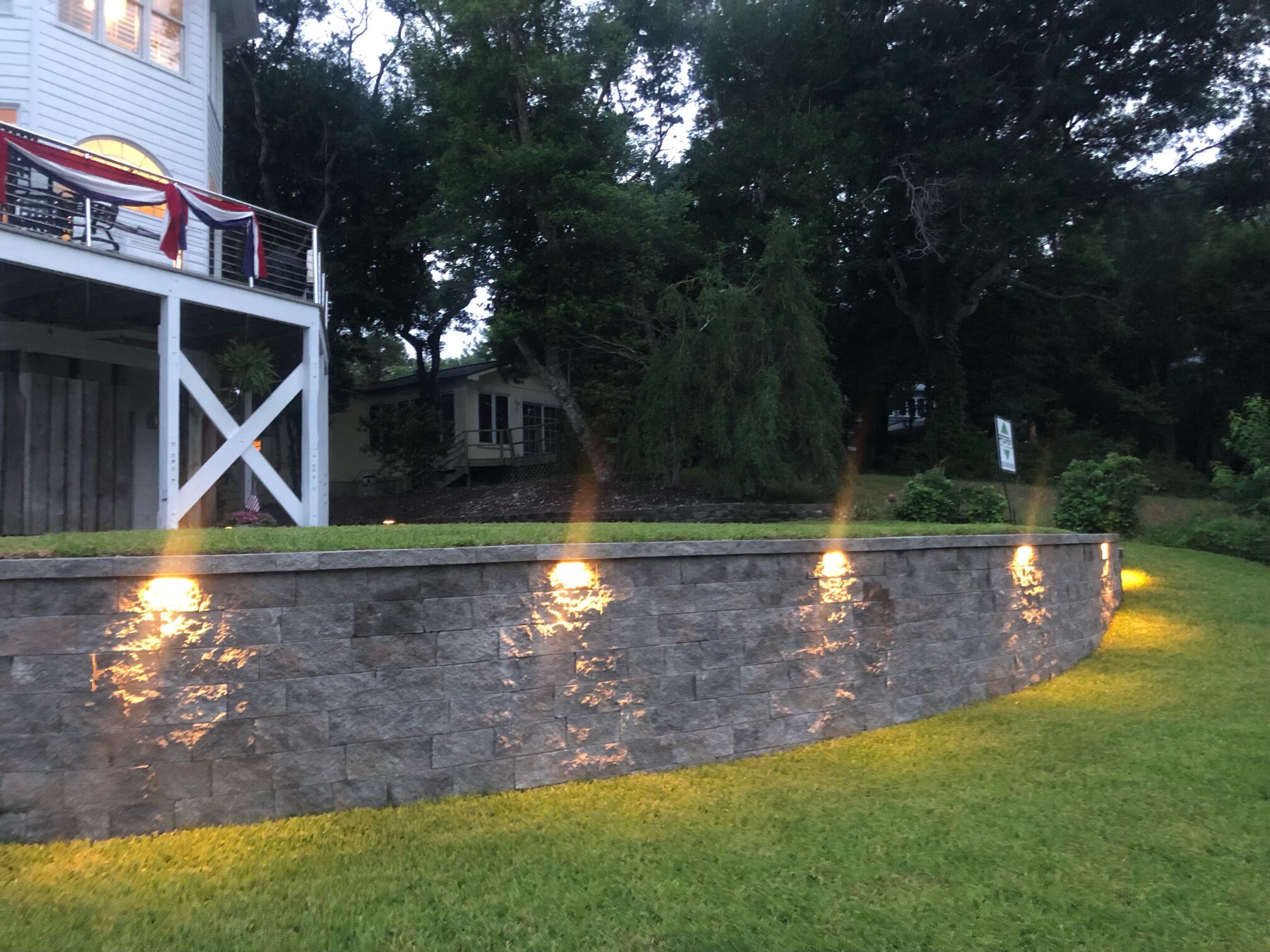House with elevated deck adorned with flags, illuminated by lights on a stone wall, set against a backdrop of lush trees and grass.