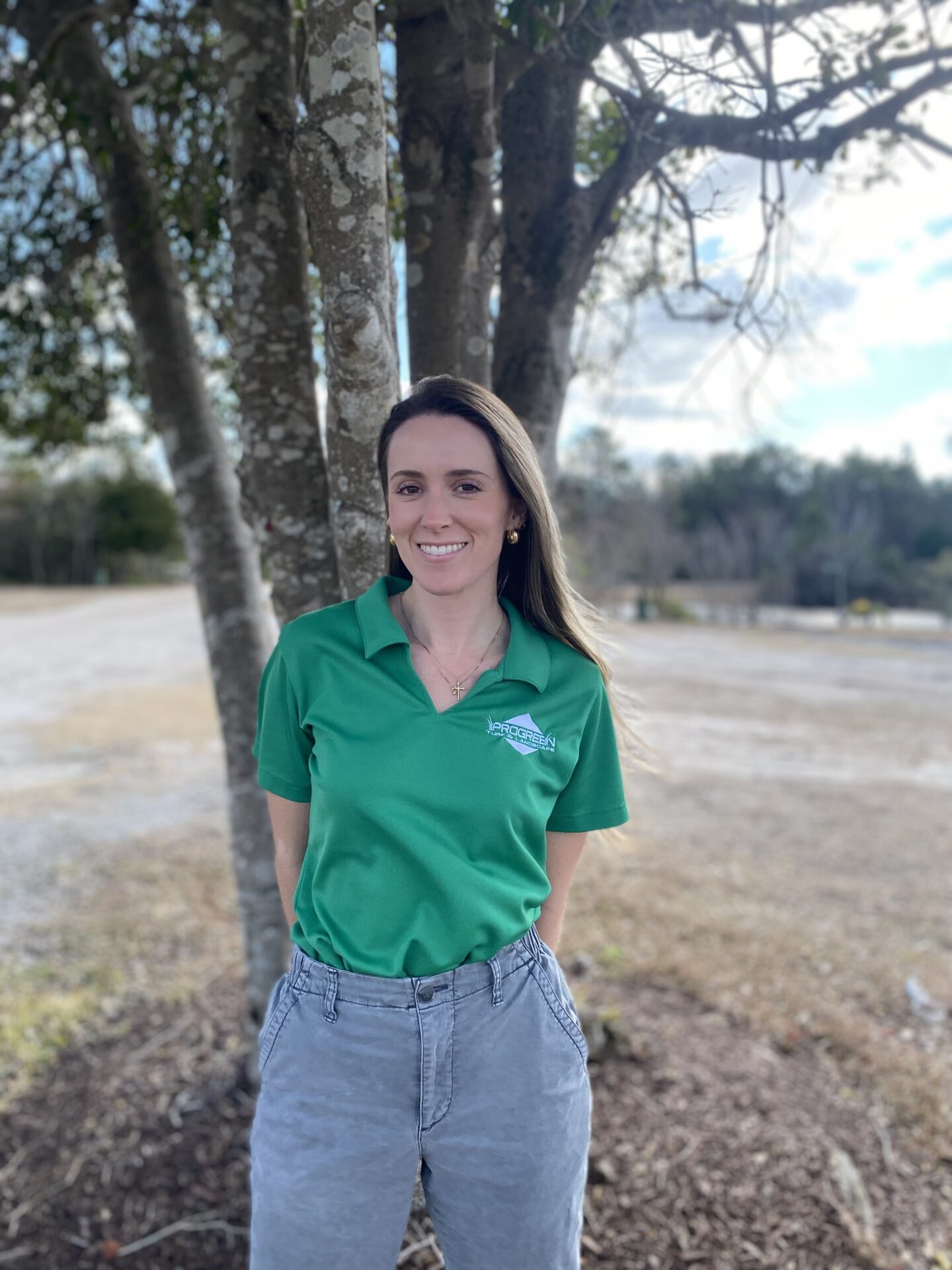 A person in a green shirt stands smiling under a tree, with a blurred outdoor background of grass and sky.