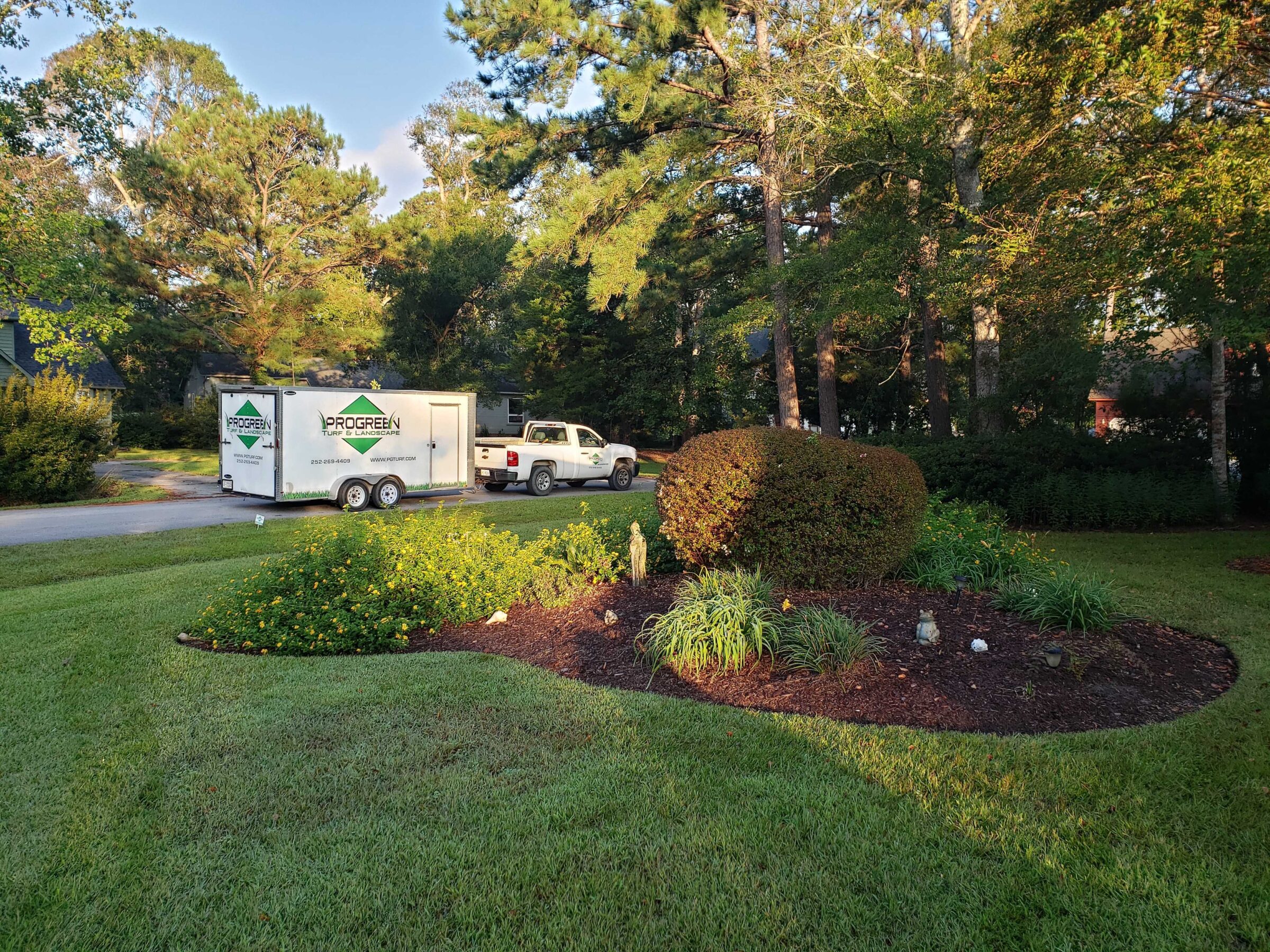 A truck with a "ProGreen Turf & Landscape" trailer is parked on a residential street, surrounded by lush trees and manicured gardens.