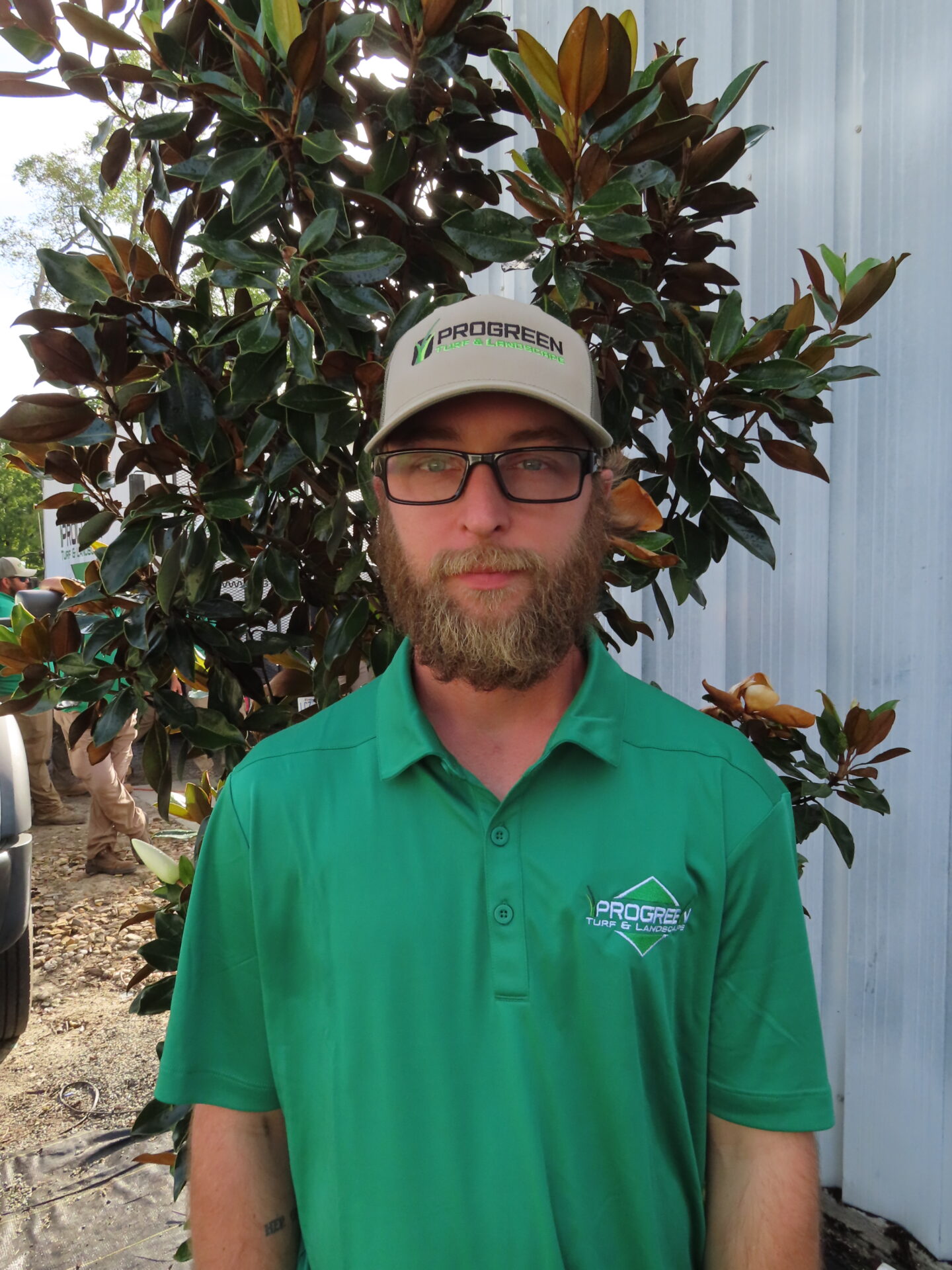 A person with a beard and glasses, wearing a ProGreen cap and shirt, stands in front of a metal building and a tree.