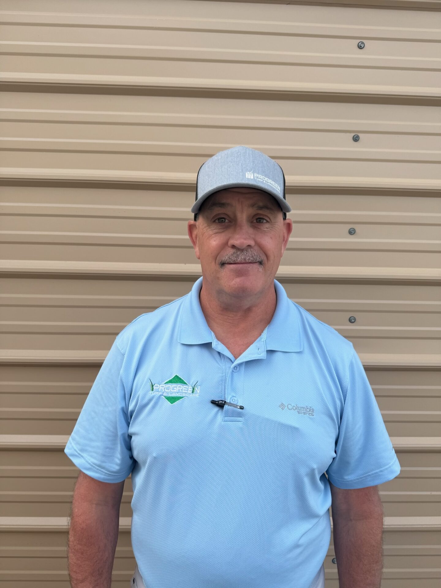 A person in a light blue shirt and cap stands against a tan corrugated metal wall, looking directly at the camera.