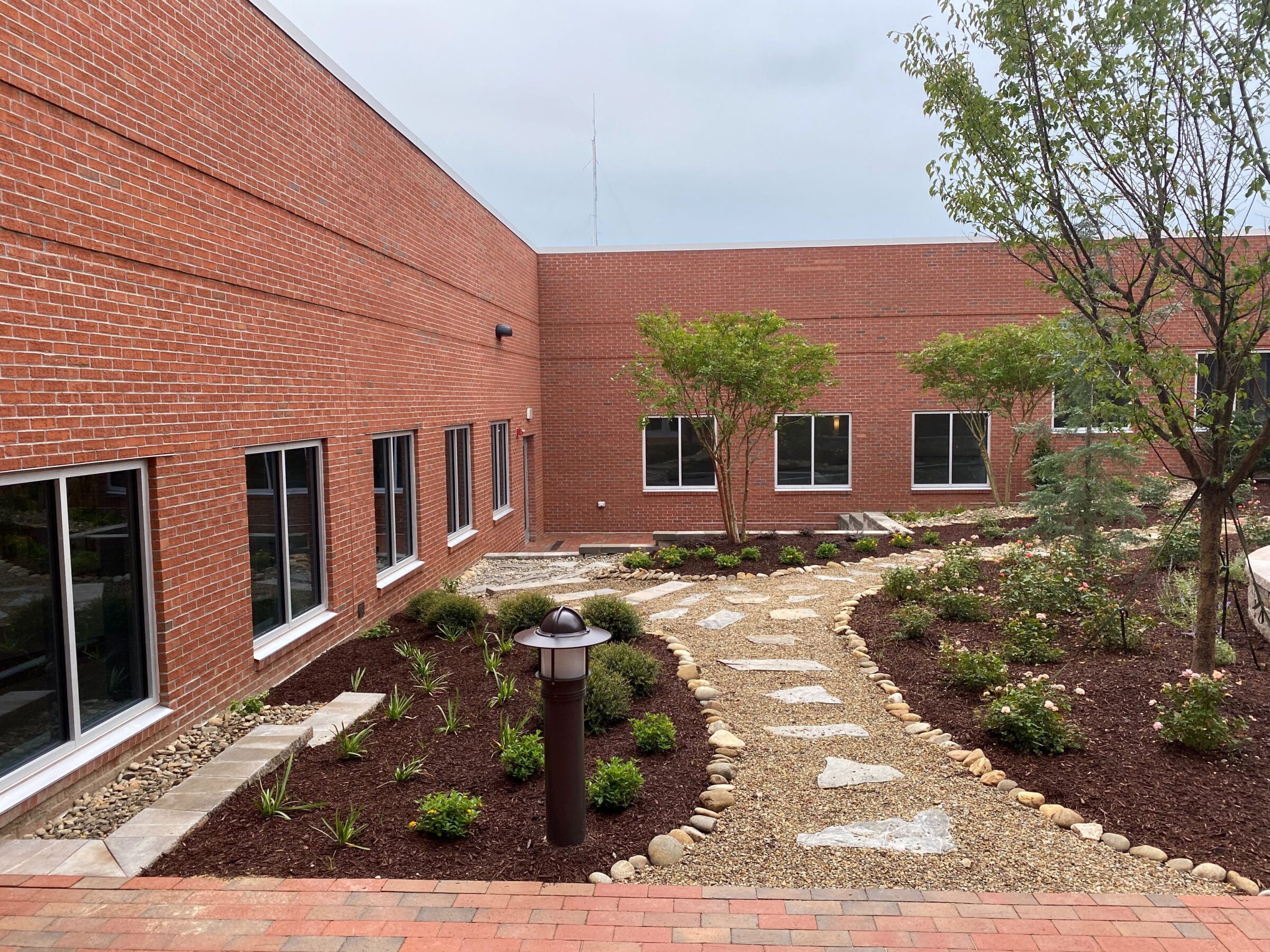A brick building courtyard with a gravel path, landscaped gardens, and trees under a cloudy sky.