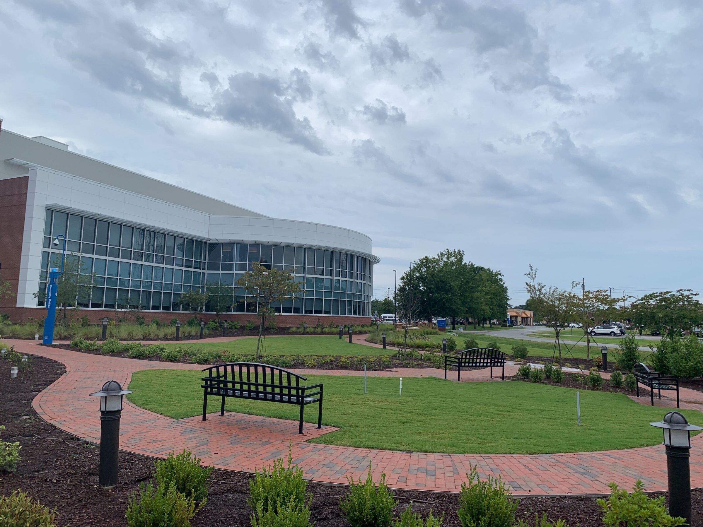 Modern building with curved glass facade, surrounded by landscaped garden with benches and brick pathways. Overcast sky and parking lot in background.