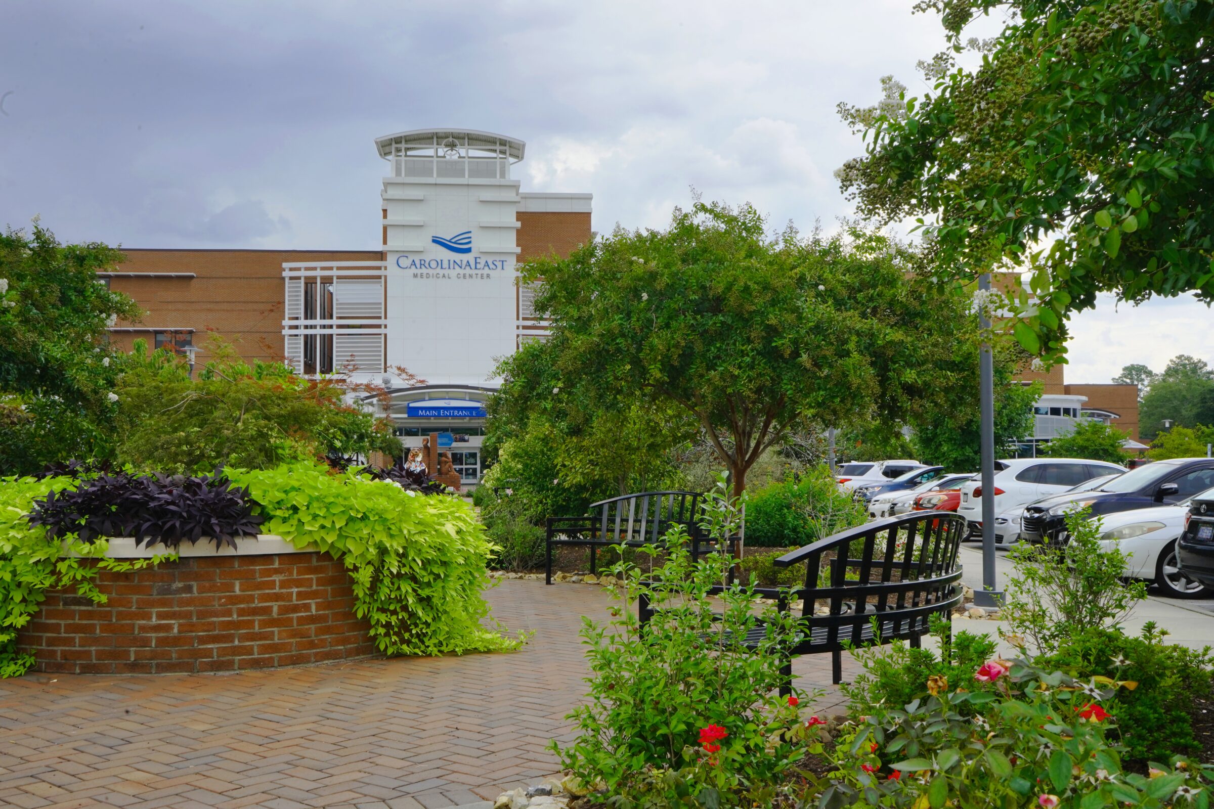 A garden outside CarolinaEast Medical Center with lush greenery, benches, and parking lot. Cloudy sky overhead provides a peaceful atmosphere.