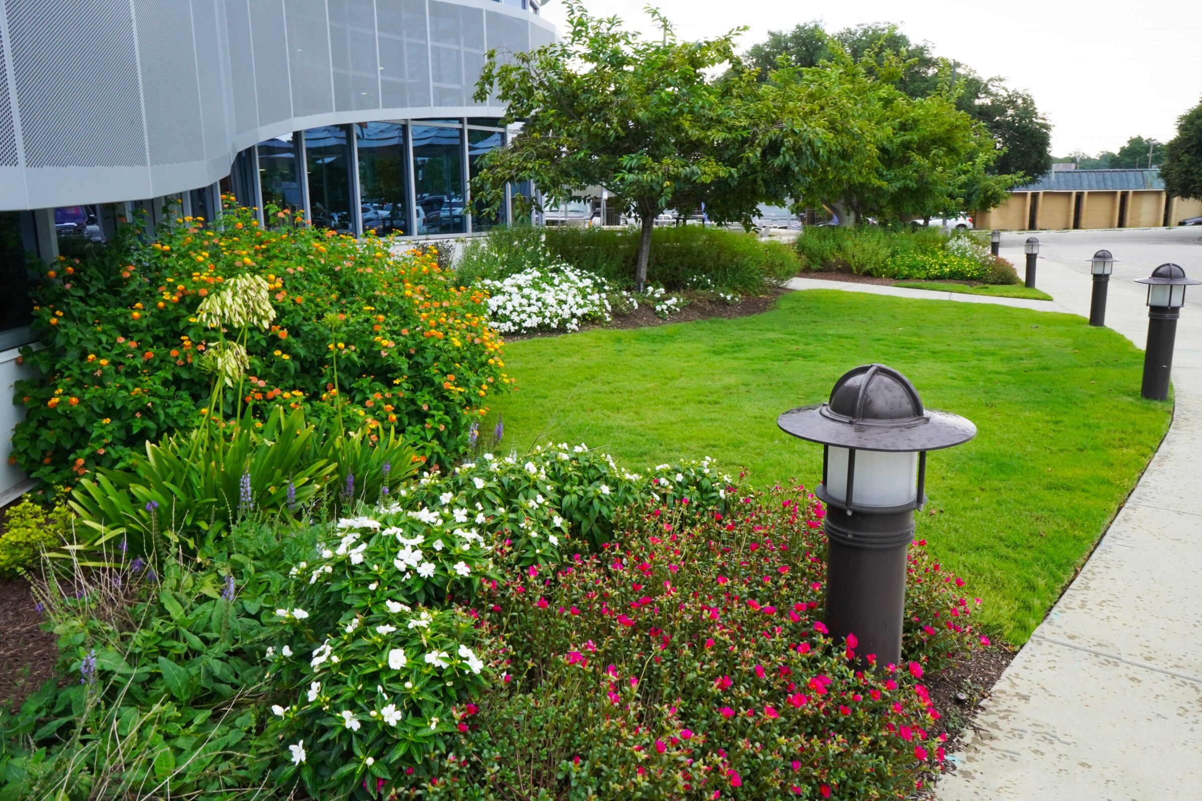 A well-maintained garden with vibrant flowers, a curved glass building, and pathway lights. Trees and a parking area are visible in the background.
