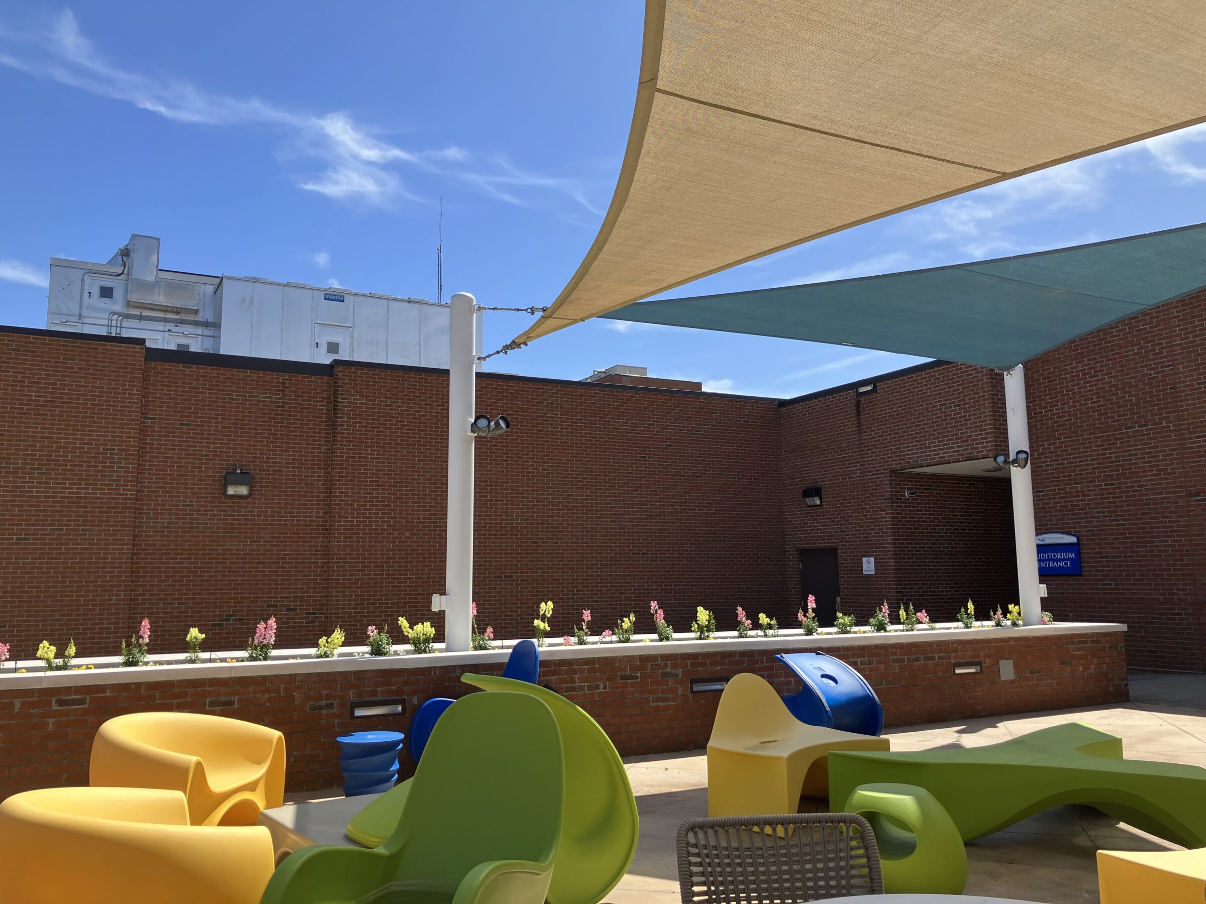 Outdoor seating area with colorful chairs and shaded sail above, surrounded by brick walls and flowers, under a blue sky.