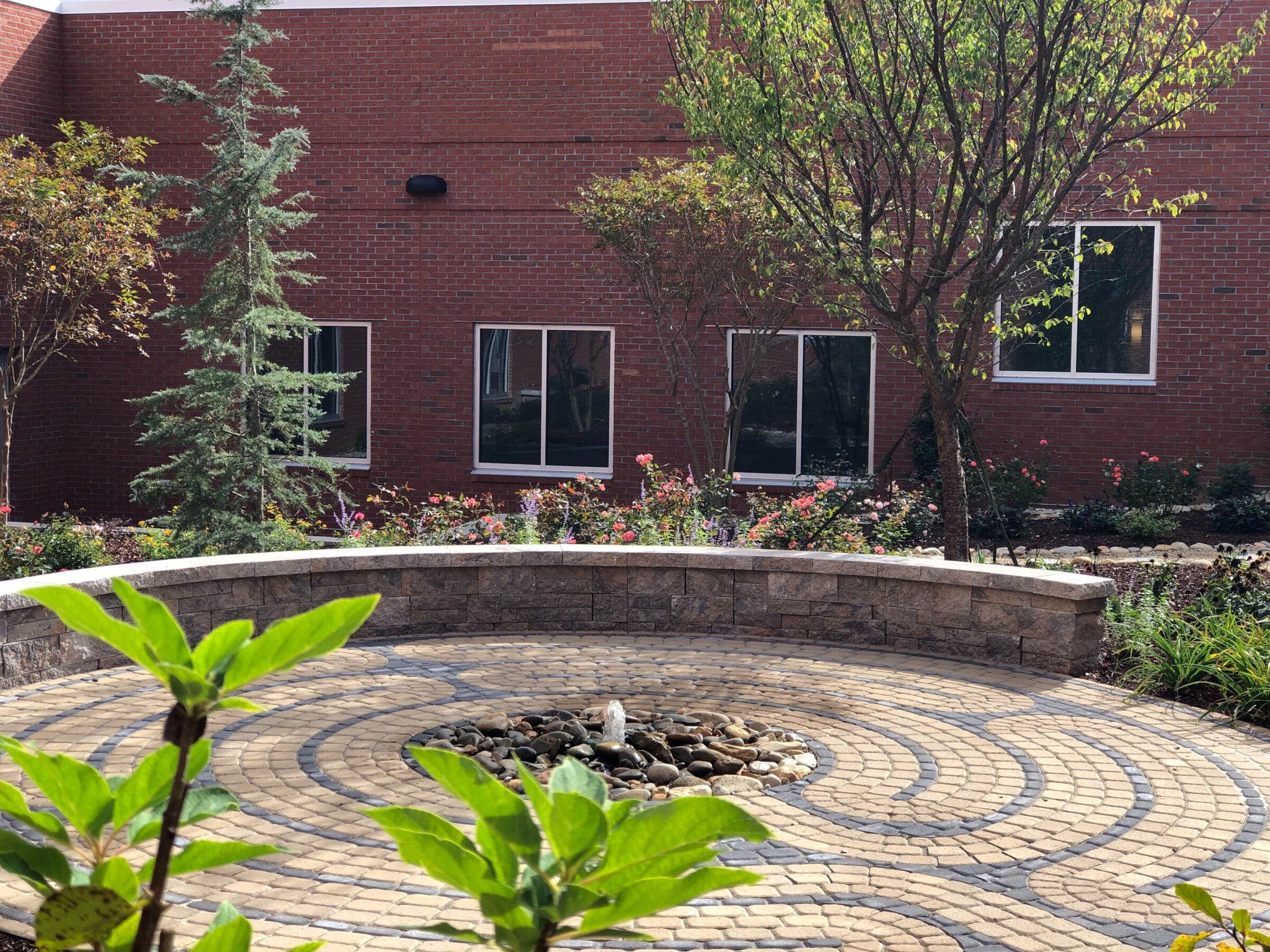 A serene courtyard with a circular stone pathway, surrounded by trees and plants, adjacent to a red brick building.