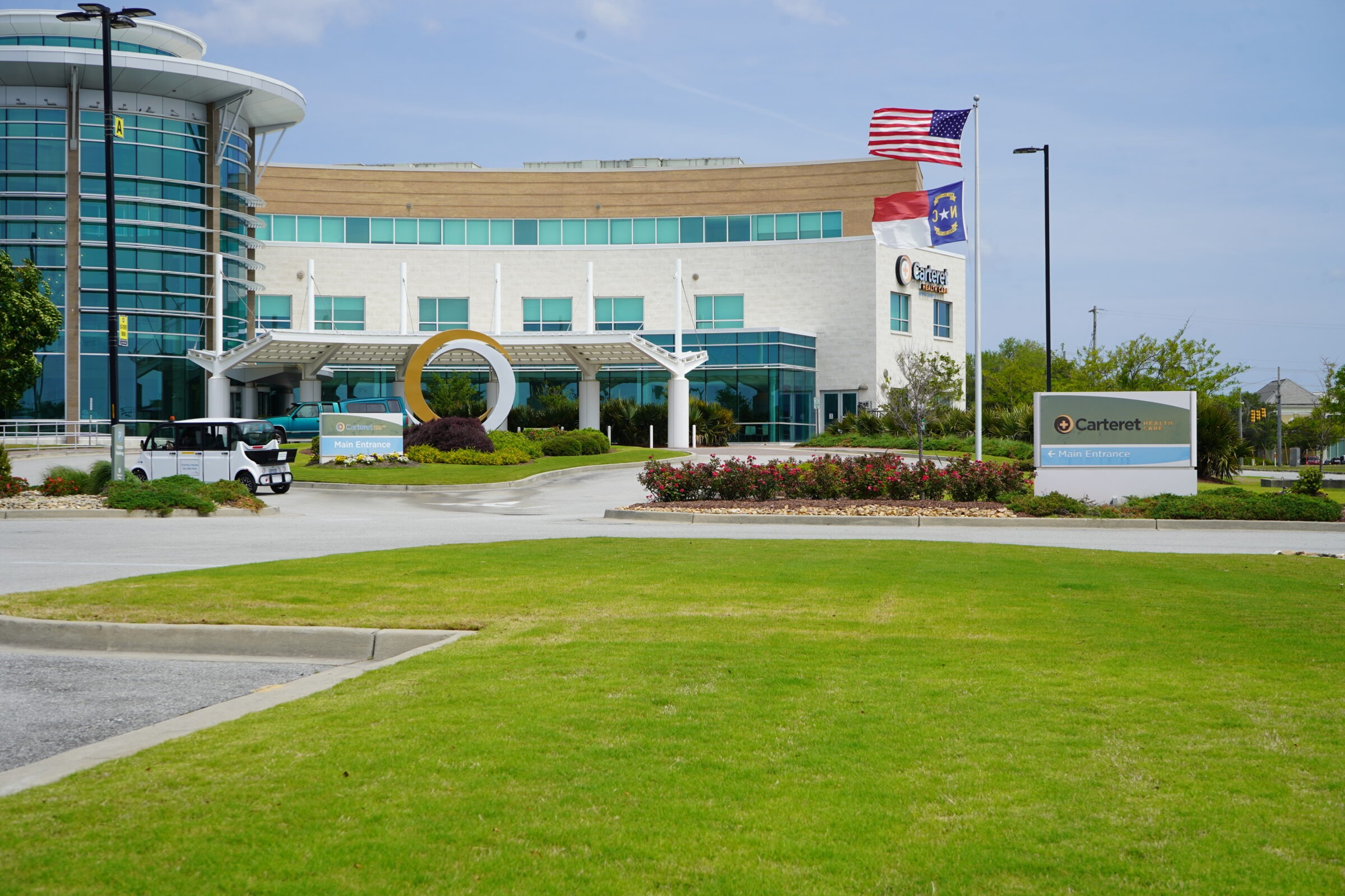 Modern building with large windows, circular sculpture, and three flags. A vehicle is parked near the entrance. Landscaping features blooming flowers and trees.