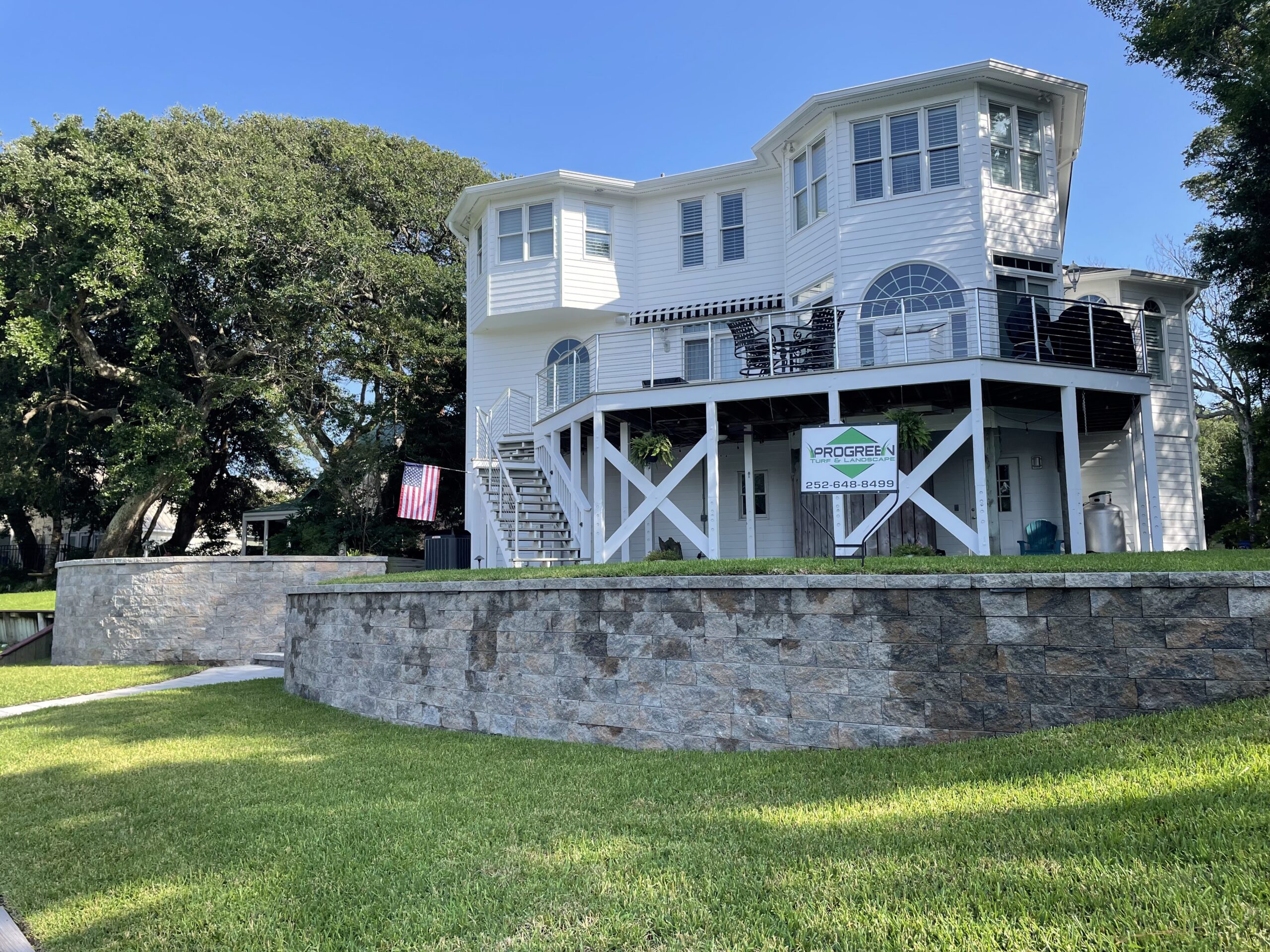 A modern, two-story white house with a spacious balcony, surrounded by lush greenery and a stone retaining wall. An American flag is displayed.
