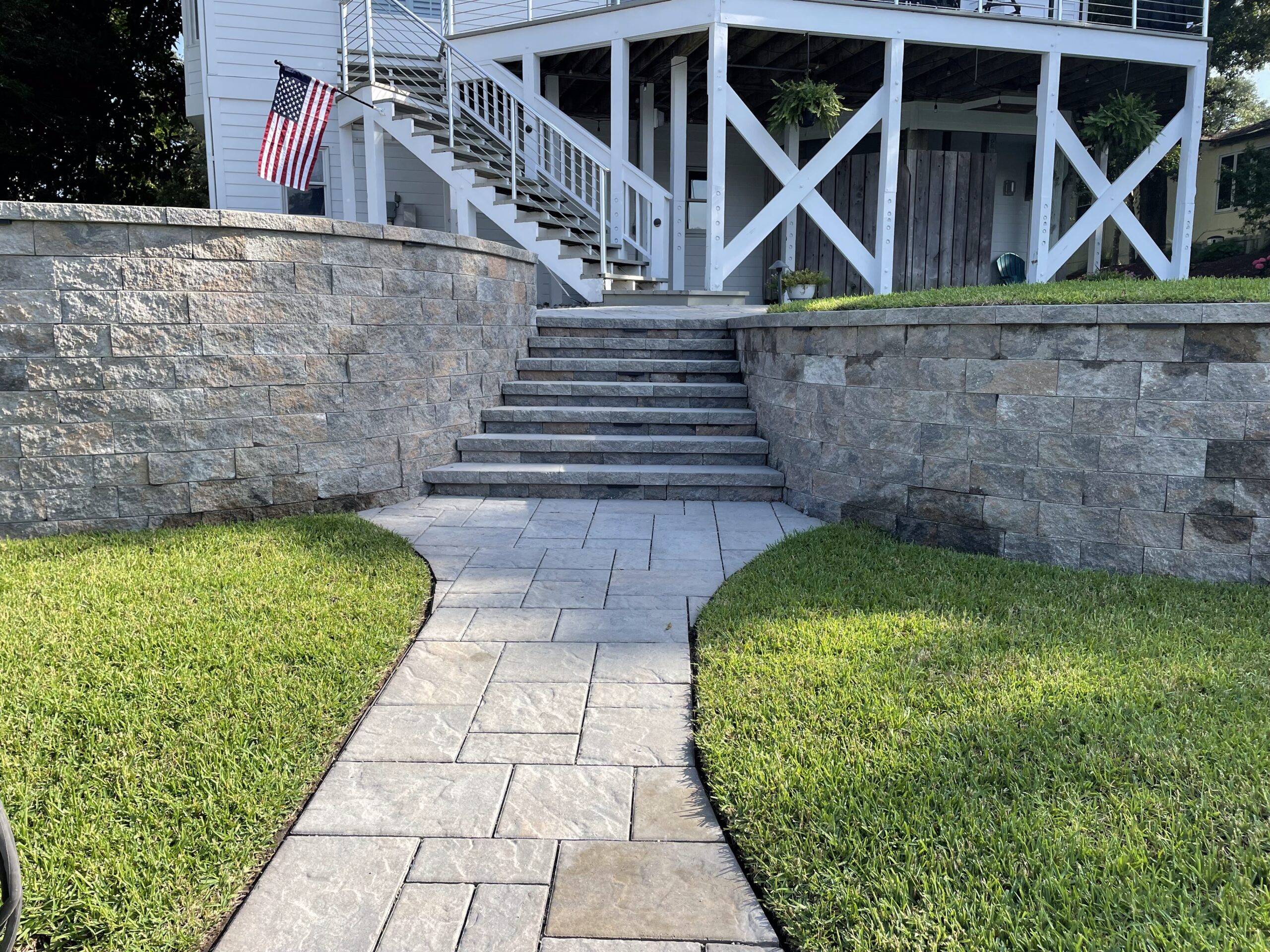 Stone pathway leads to elevated porch with American flag, surrounded by neatly trimmed grass and stone retaining walls. Modern railings and ferns decorate.