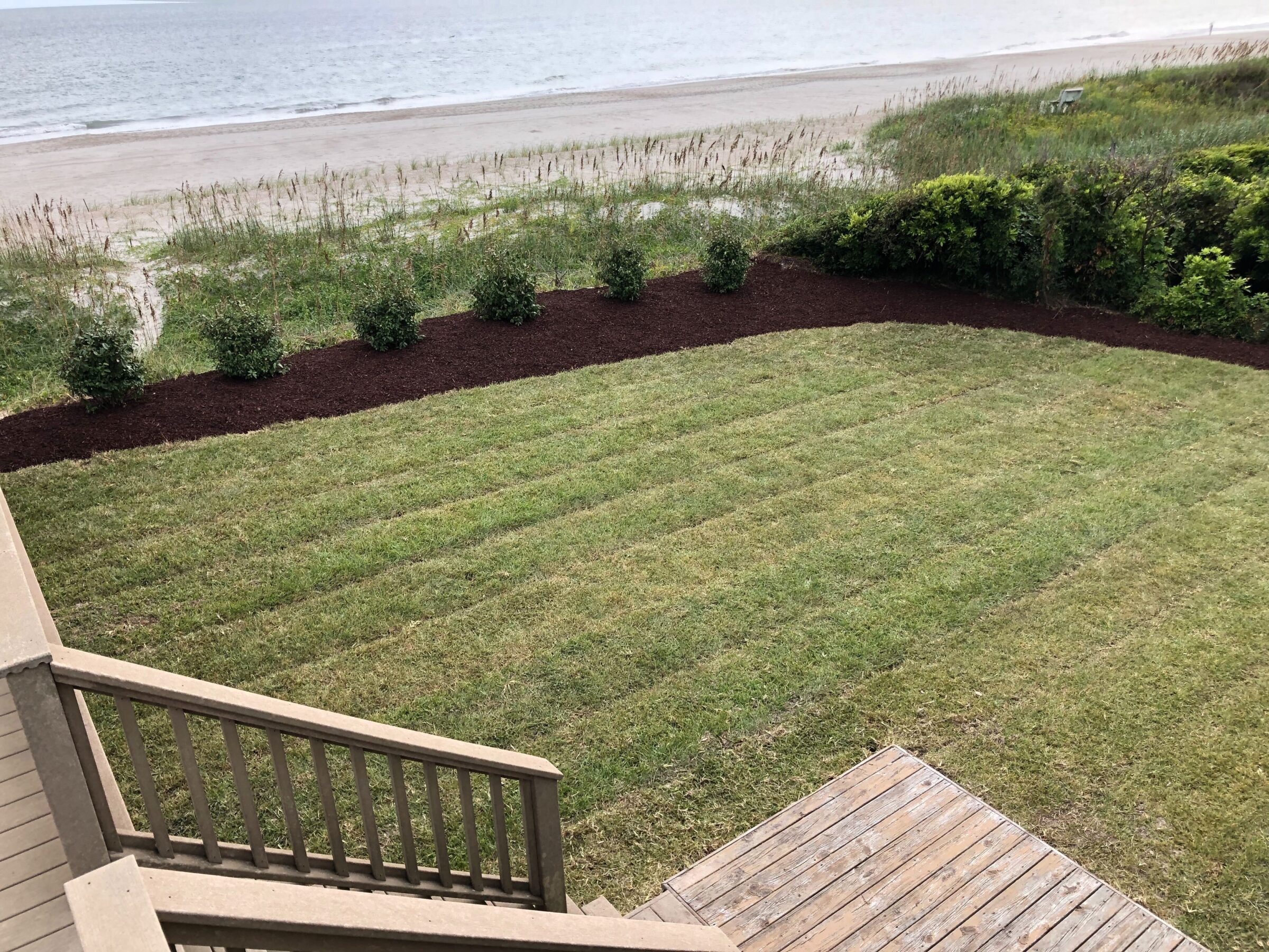 A wooden deck overlooks a neatly trimmed lawn with shrubs, leading to a sandy beach and calm ocean waves under a cloudy sky.