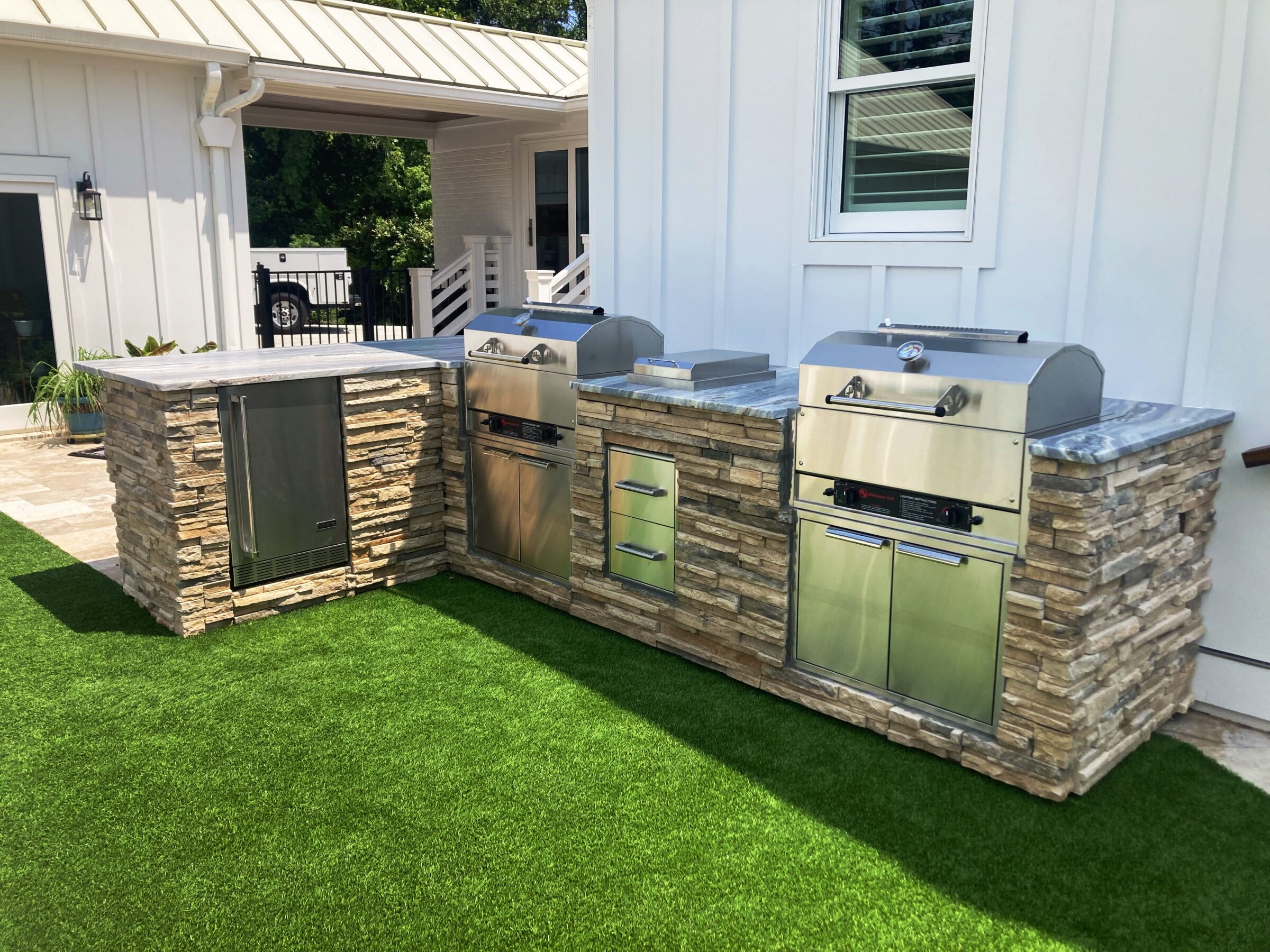 Outdoor kitchen with stone countertops, built-in grills, and stainless steel appliances on artificial grass beside a modern white house with a metal roof.