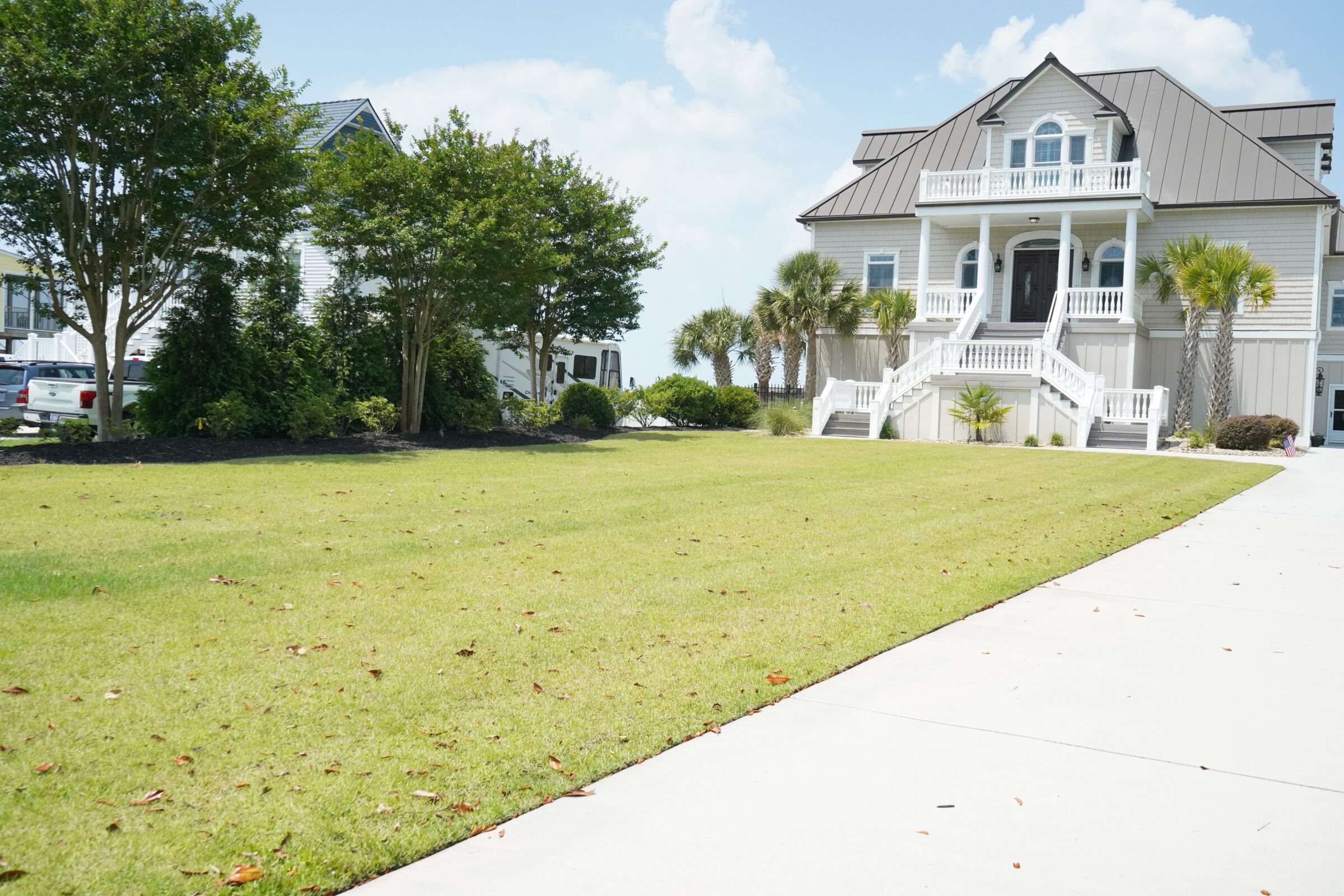 A large, elegant house with a spacious, manicured lawn and palm trees. The driveway extends toward the entrance, lined with greenery.