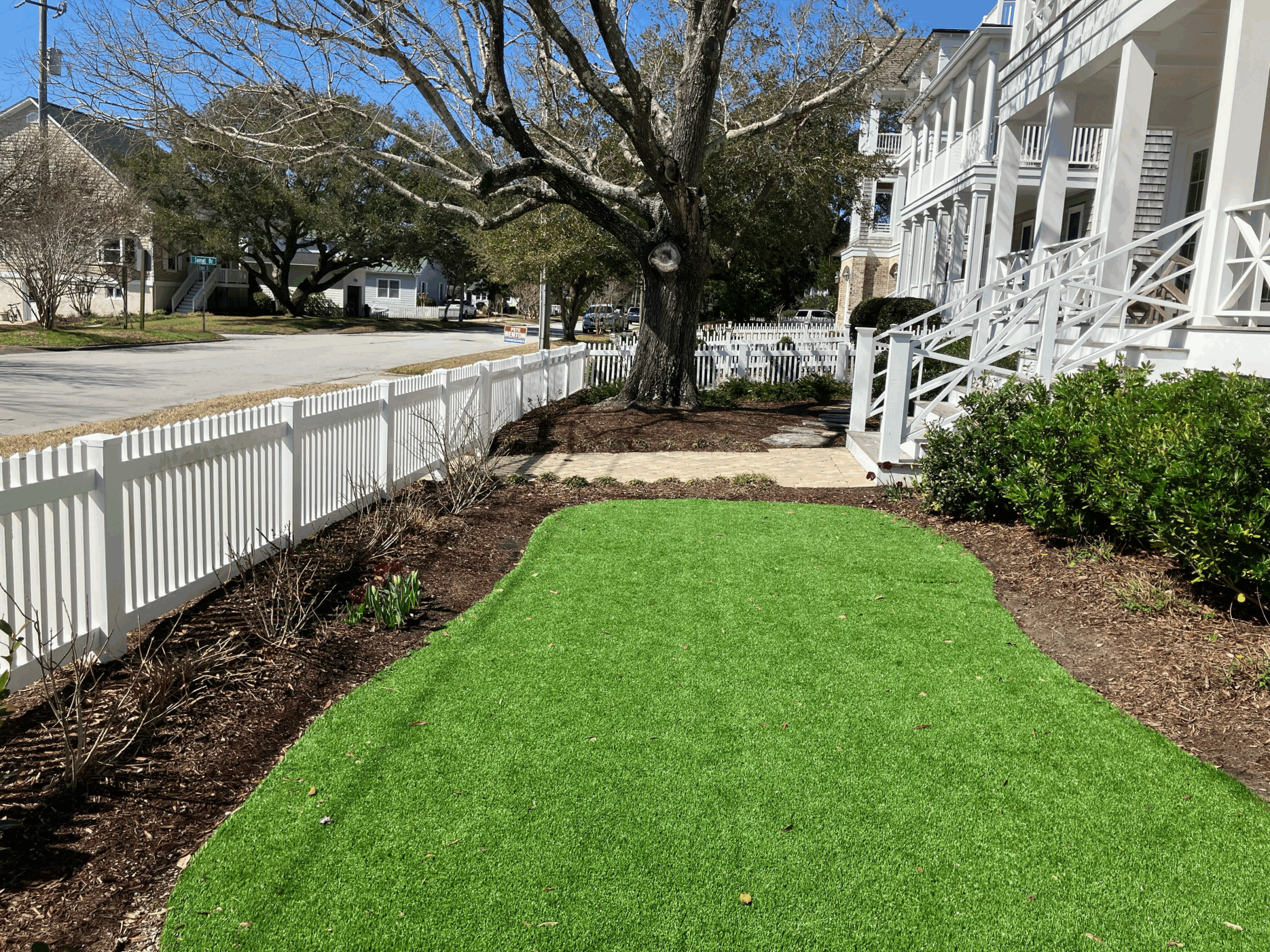 White picket fence, green lawn, bare trees, and white house with columns create a picturesque neighborhood scene under a clear blue sky.