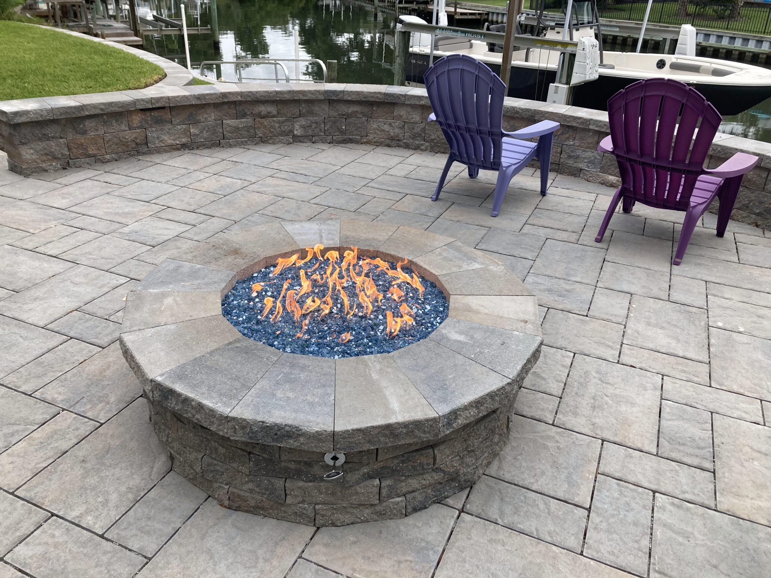 A fire pit with blue glass and flames, surrounded by stone, overlooks a canal with purple chairs and boats nearby.