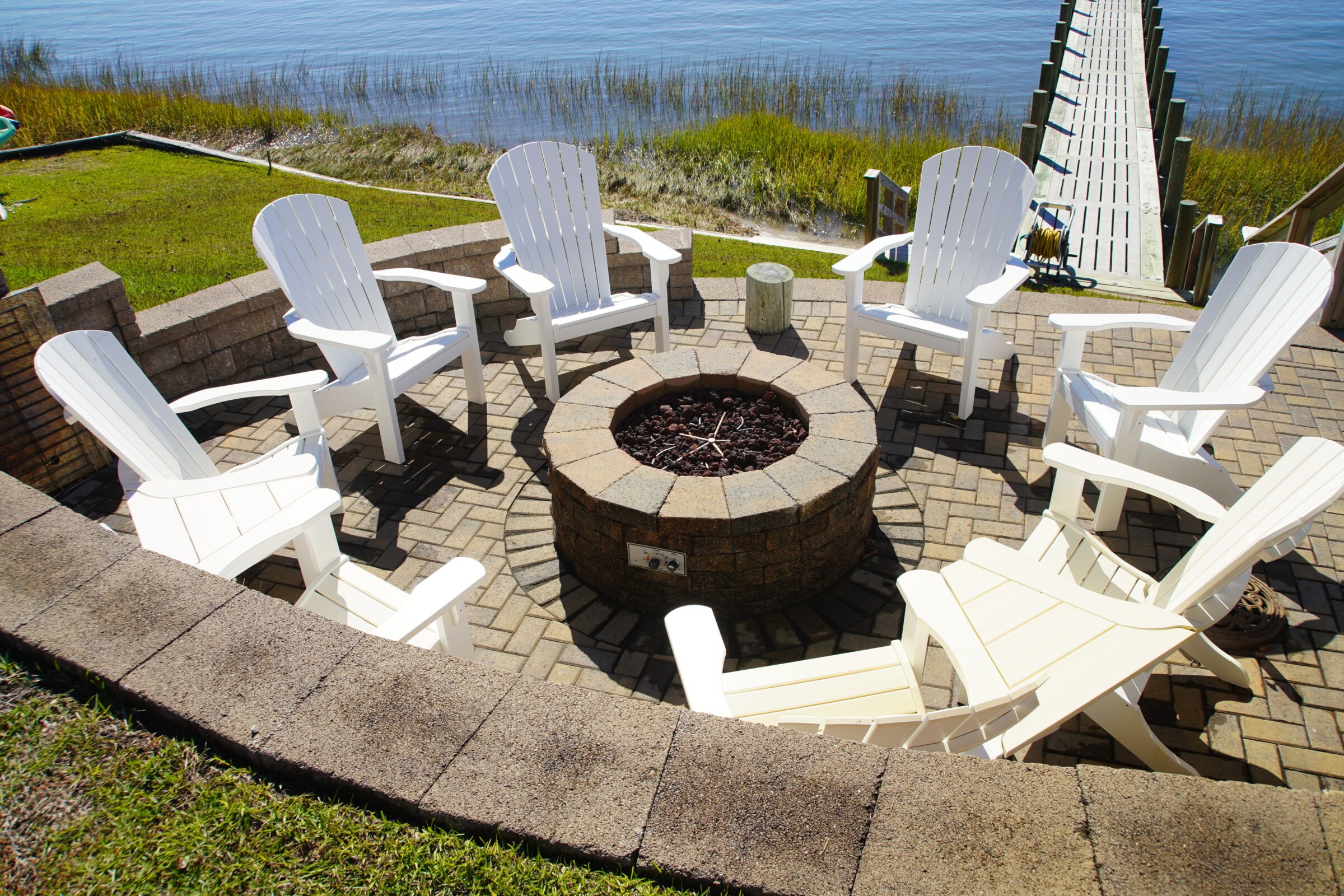 A wooden dock extends over a lake, bordered by grass, with white Adirondack chairs surrounding a stone fire pit on the patio.