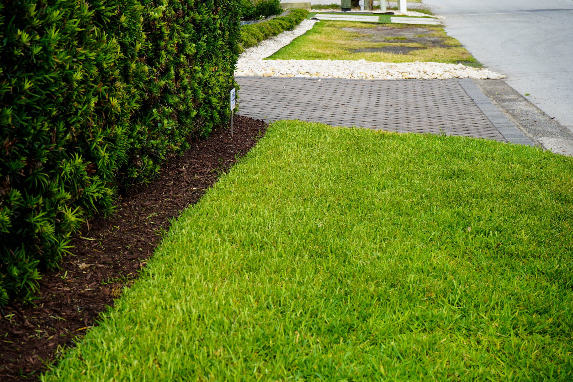 A well-maintained lawn and trimmed hedge line a brick-paved walkway, with a gravel border and street visible in the background.