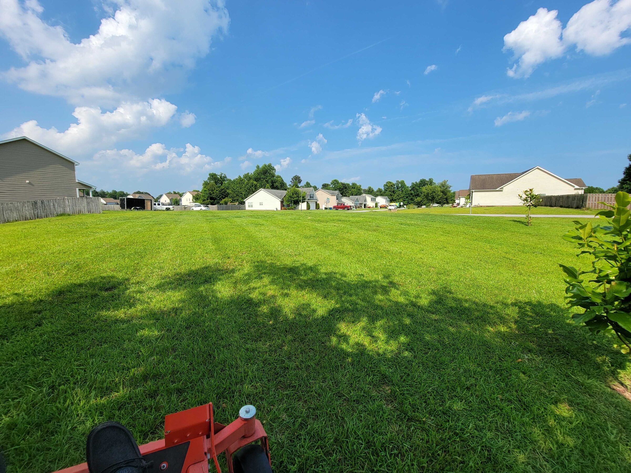 A sunny neighborhood scene featuring a large green lawn, houses, and a clear blue sky with scattered clouds. No landmarks visible.