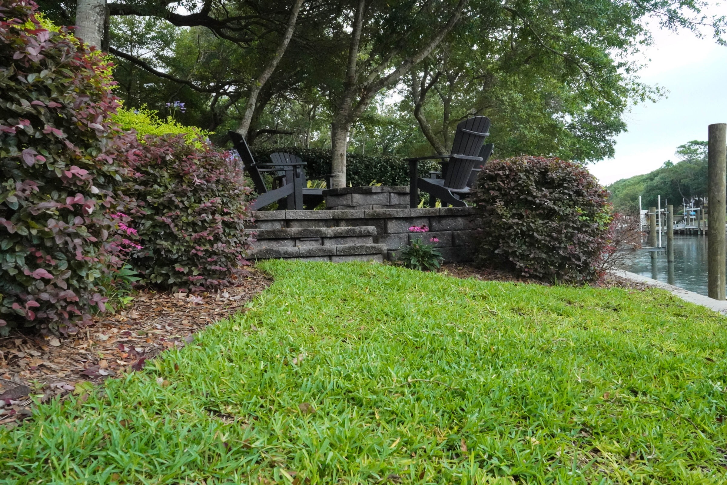 A serene garden scene features Adirondack chairs, lush greenery, and a waterside area with docks, creating a peaceful outdoor setting.