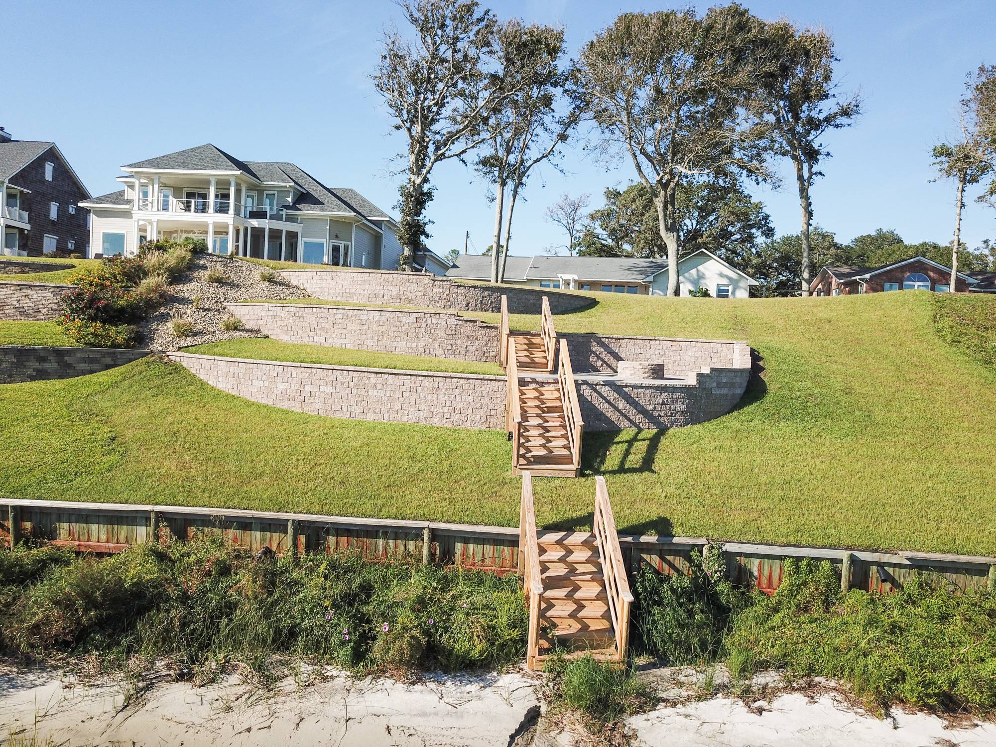 A coastal home with tiered landscaping, wooden stairs, and lush greenery. Sunny day with clear blue skies and a distant sand area.
