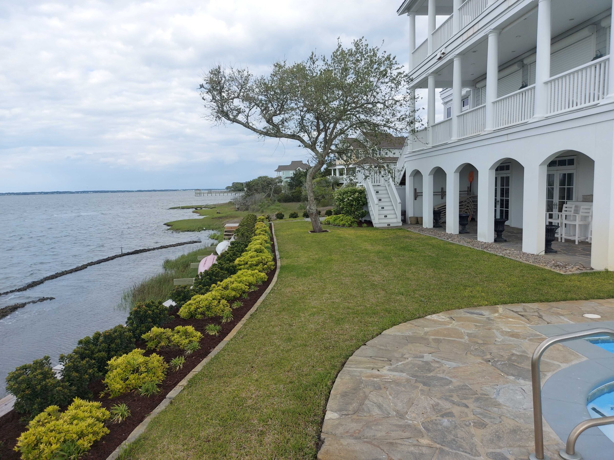 A coastal house with white columns faces a calm body of water. Well-manicured lawn with hedges lines the shoreline, under a cloudy sky.