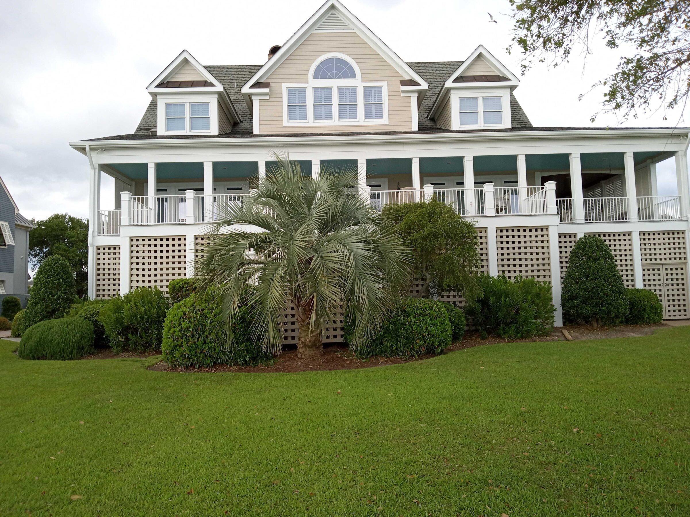 Spacious two-story house with attic, large porch, and manicured garden. Features palm tree and white latticework. Overcast sky above.