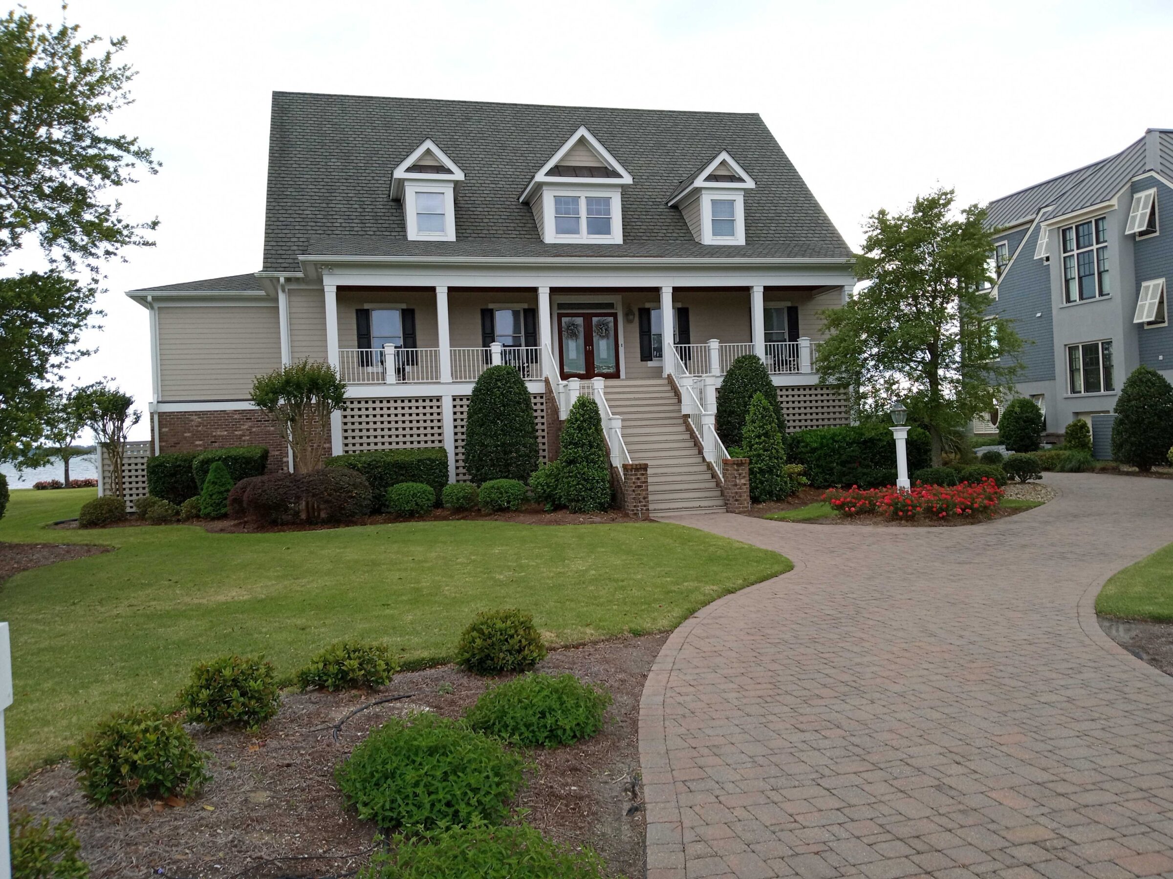 A two-story house with a gabled roof, surrounded by a neat garden and pathway, stands adjacent to another gray building, trees nearby.
