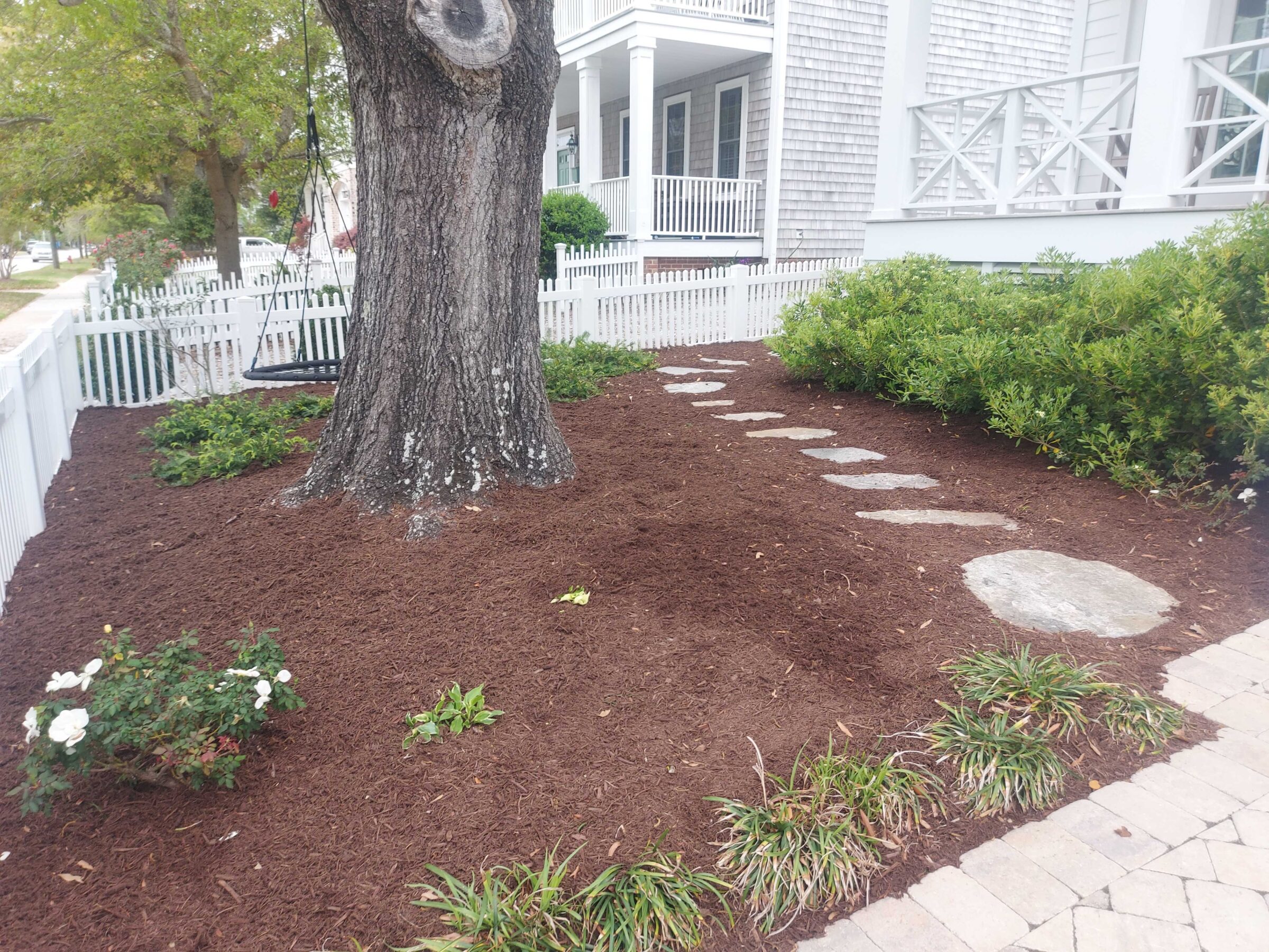 A neatly landscaped garden features a tree with a swing, stepping stones, and a white picket fence surrounding a porch.