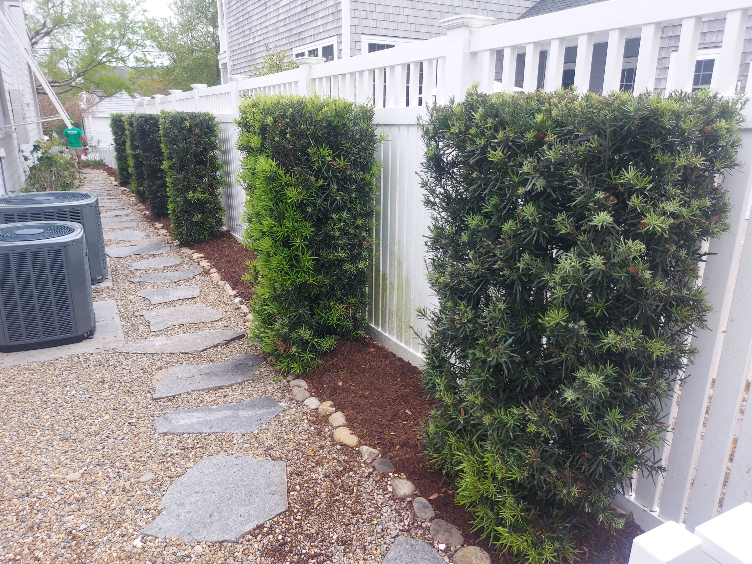 Stone path lined with neatly trimmed bushes beside a white fence. Visible air conditioning units positioned on the left. Person walks in the distance.