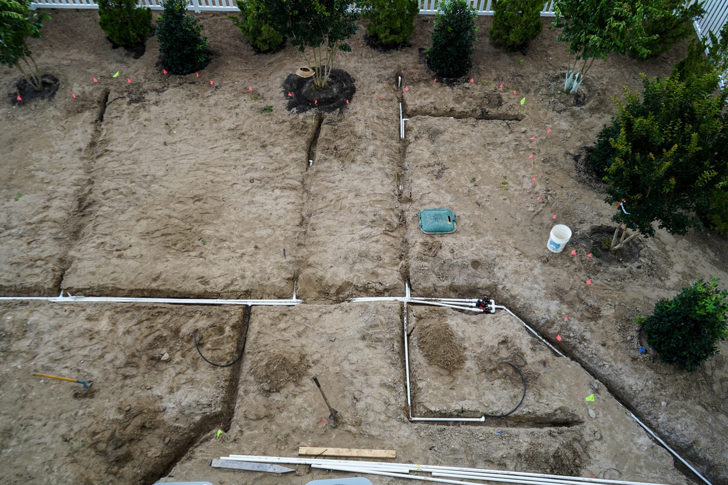 A yard under construction with exposed pipes, trenches, and newly planted trees. White fence and tools are visible in the background.