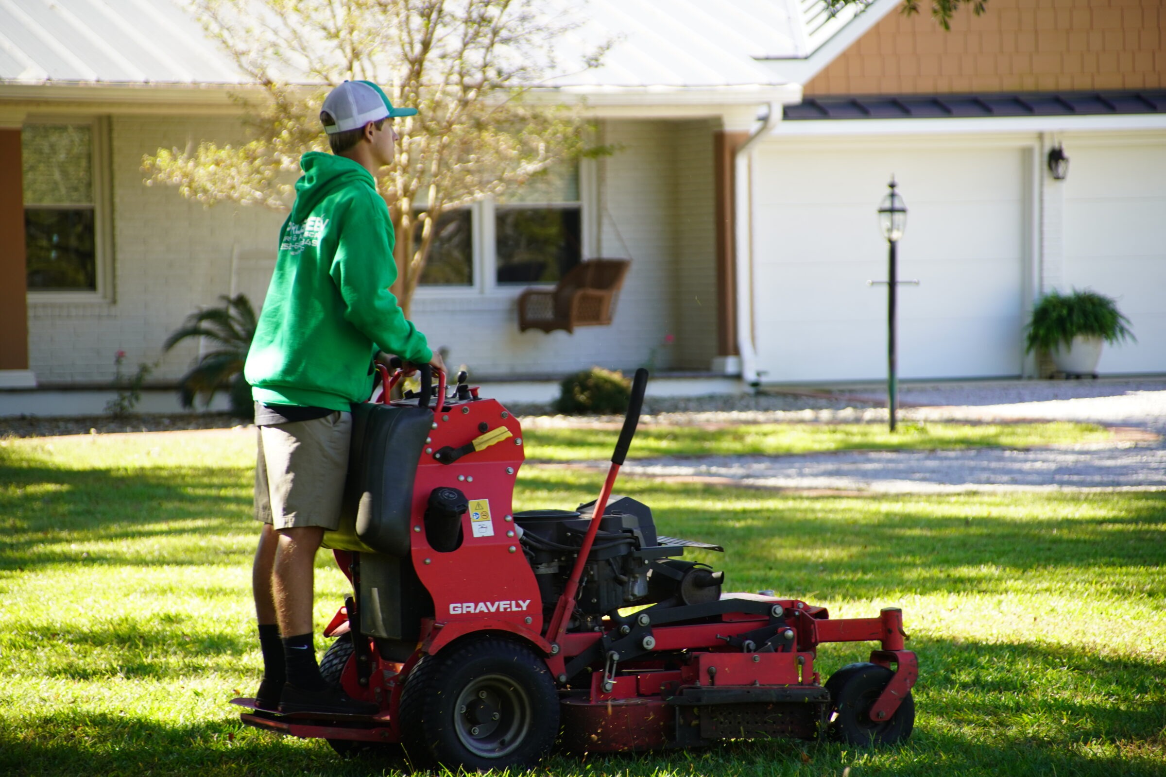 A person operating a lawnmower on grassy front yard, wearing a green hoodie and cap, near a residential house and a small mailbox.
