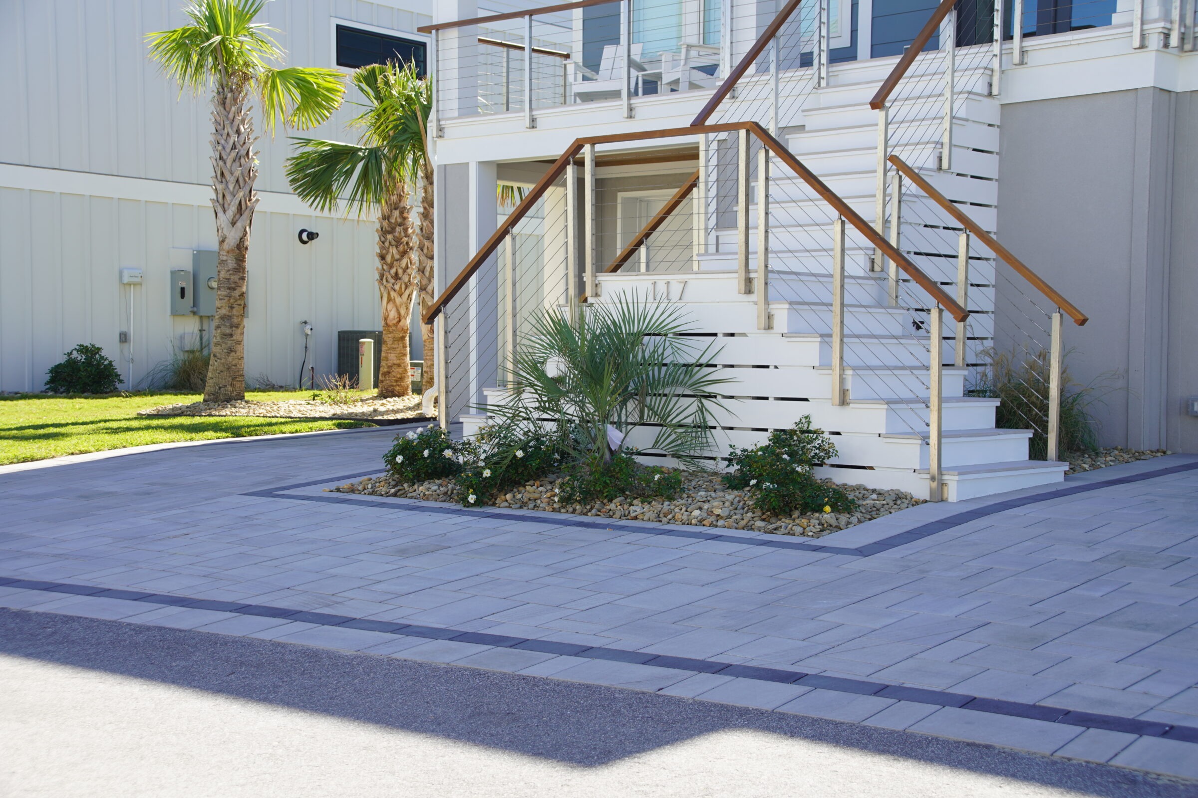 Modern house exterior with white steps, sleek railings, palm trees, and landscaped garden. Paved driveway enhances the clean, coastal look. No people visible.