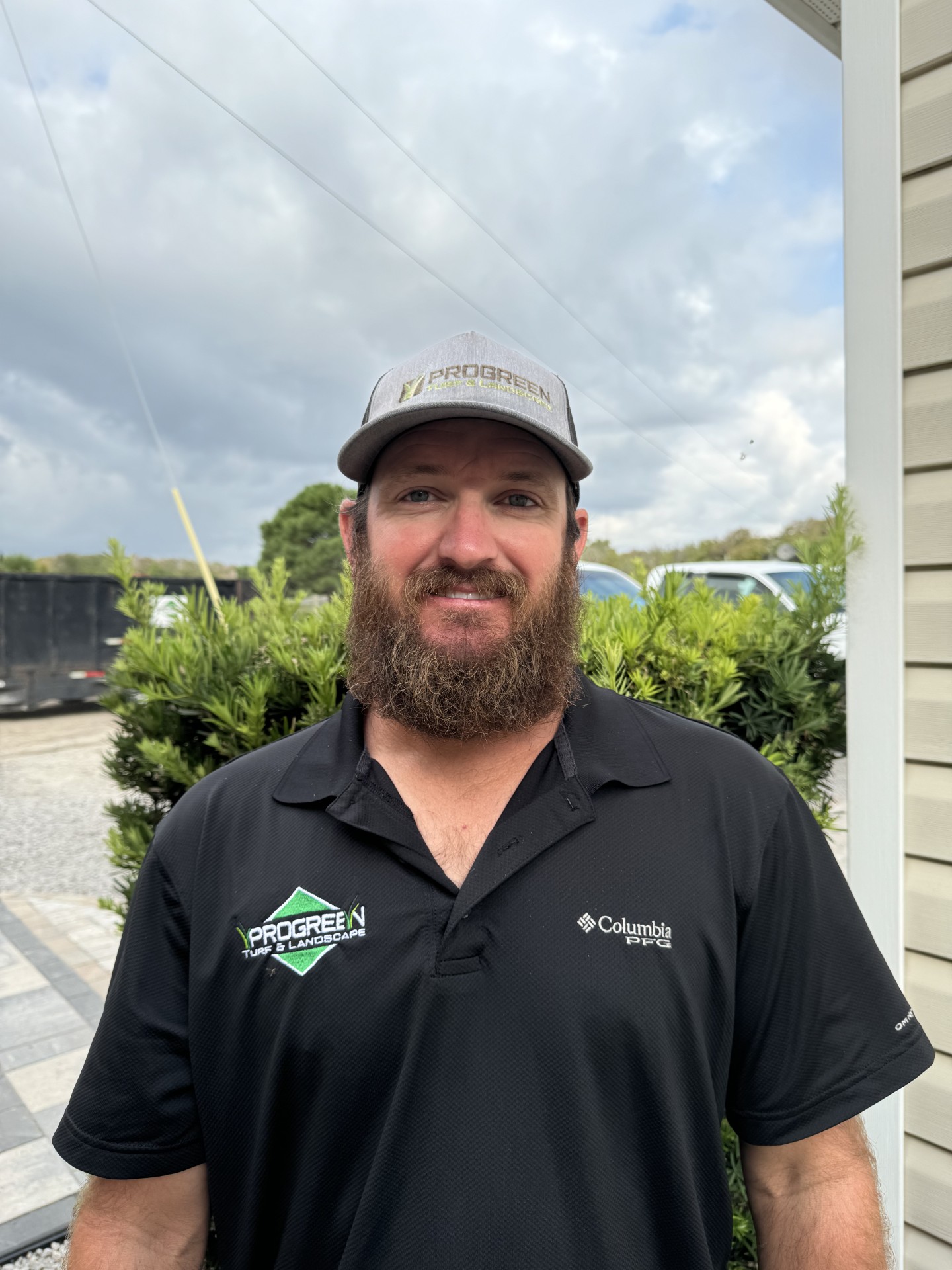 A person wearing a "ProGreen" cap and black shirt stands outdoors, smiling. Cloudy sky and greenery in the background.