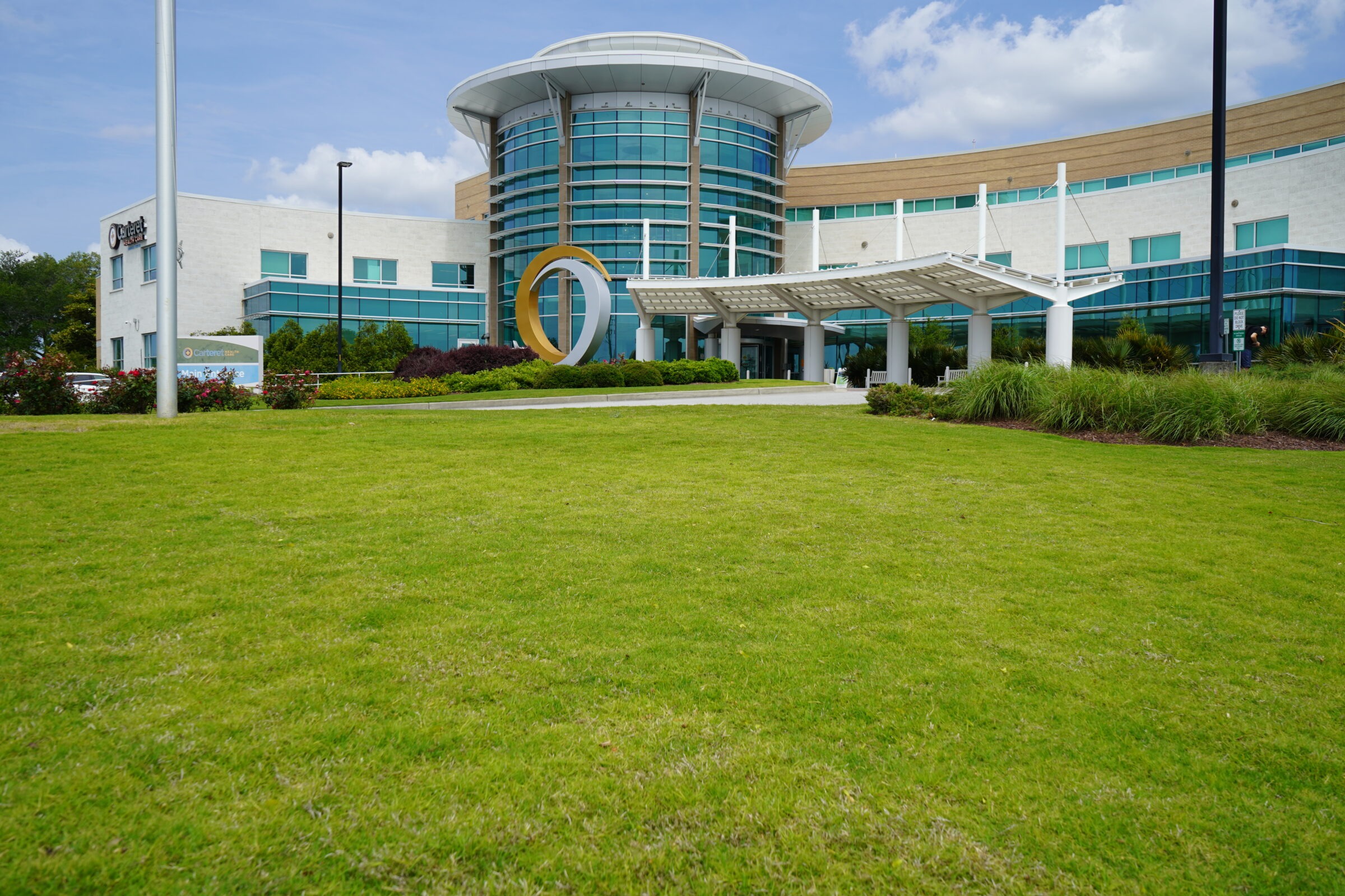 Modern glass building with circular entrance design and green lawn. Overcast sky. Two people nearby. Hospital structure, no recognizable landmarks or historical buildings.