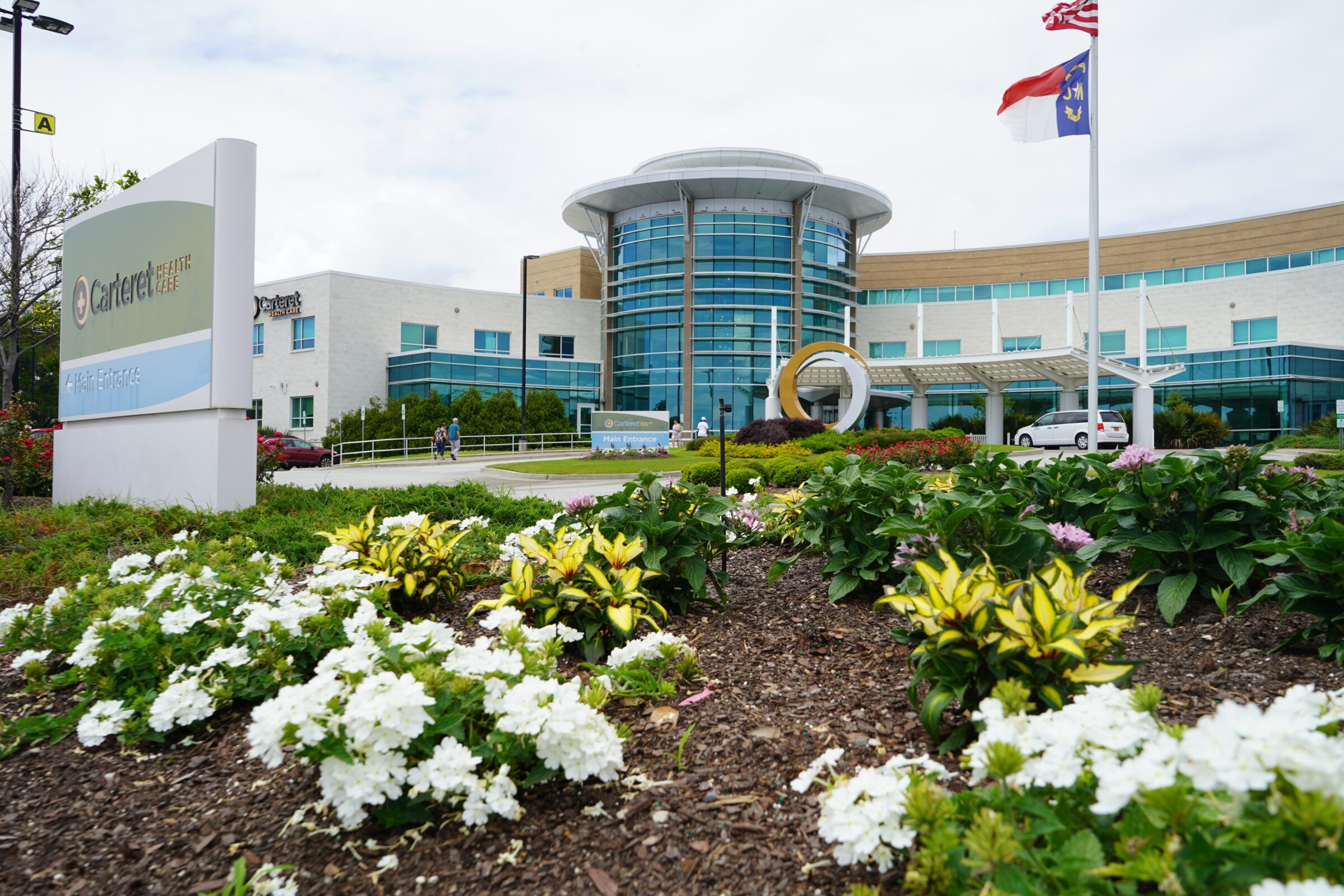 Modern building with glass facade, landscaped garden, and Carteret Health Care sign. Two flags and a few people are visible on the premises.