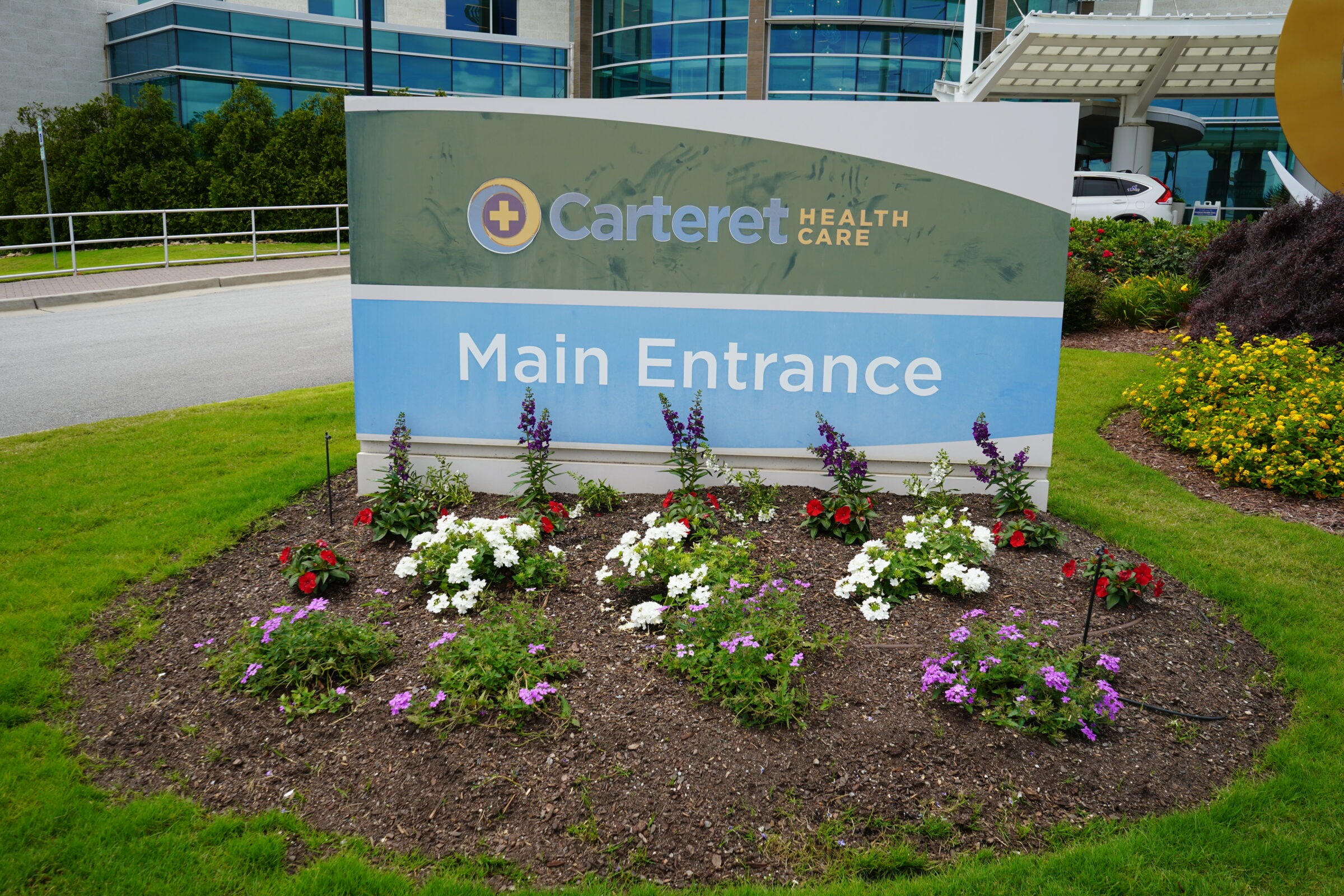Signage for Carteret Health Care's Main Entrance, surrounded by colorful flowers and greenery, near a modern building with glass windows.