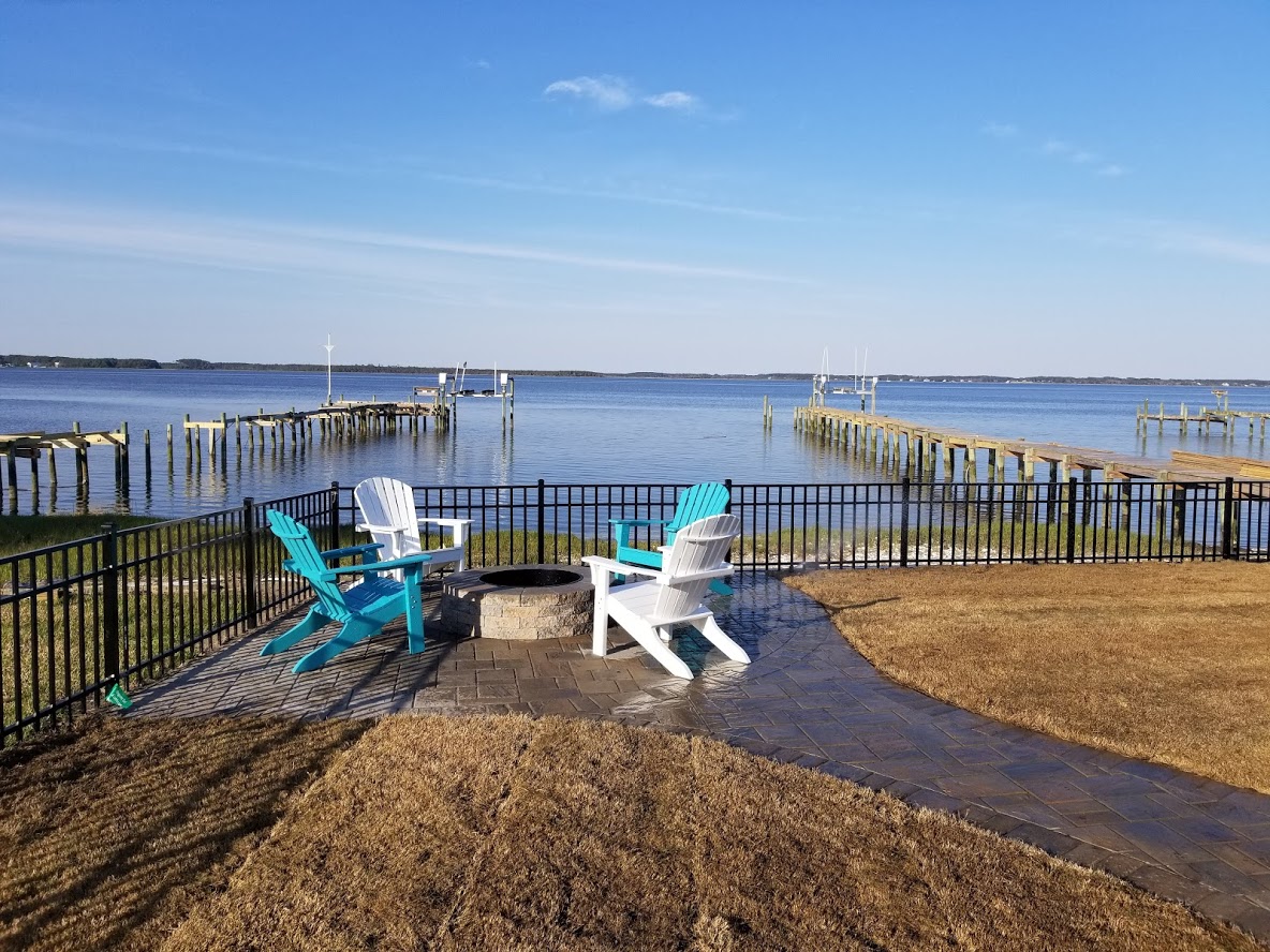 Two Adirondack chairs face a waterfront view with docks, enclosed by a black fence on a sunny day.