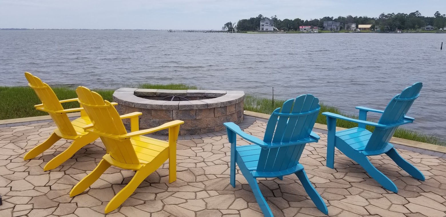 Four colorful Adirondack chairs surround a stone fire pit on a patio, overlooking a tranquil lake with houses lined along the distant shore.