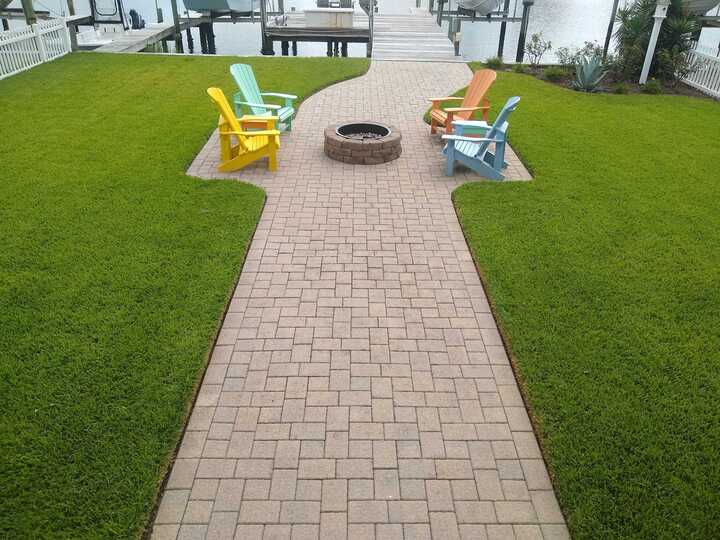 A patio with colorful chairs around a fire pit, overlooking a boardwalk leading to a tranquil waterfront. Lush green lawn surrounds the scene.