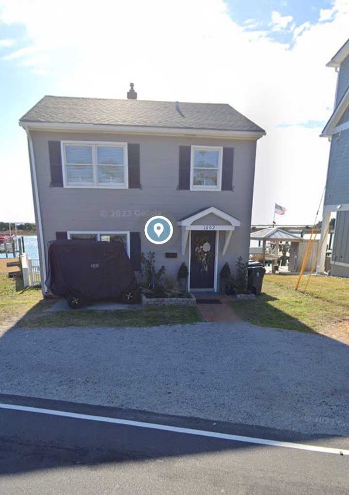 Two-story gray house with black shutters, American flag, driveway, parked car, and front garden, located beside a body of water on sunny day.