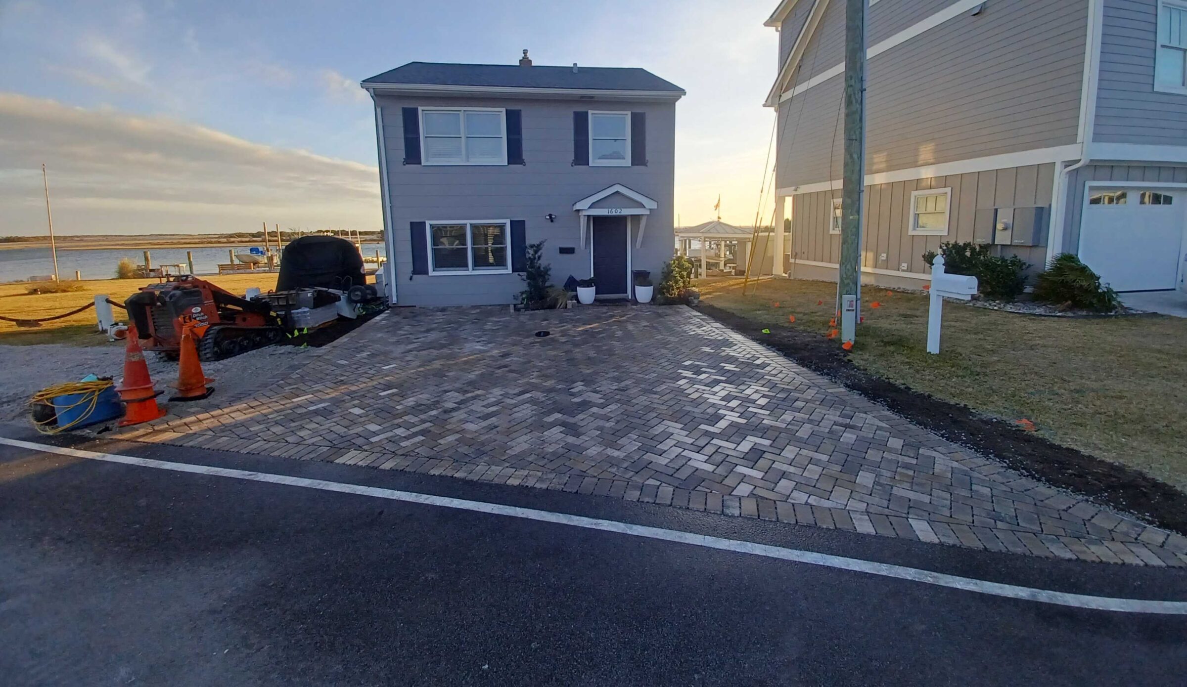 A two-story house with a brick driveway is near water and construction equipment, under a partly cloudy sky and setting sun.