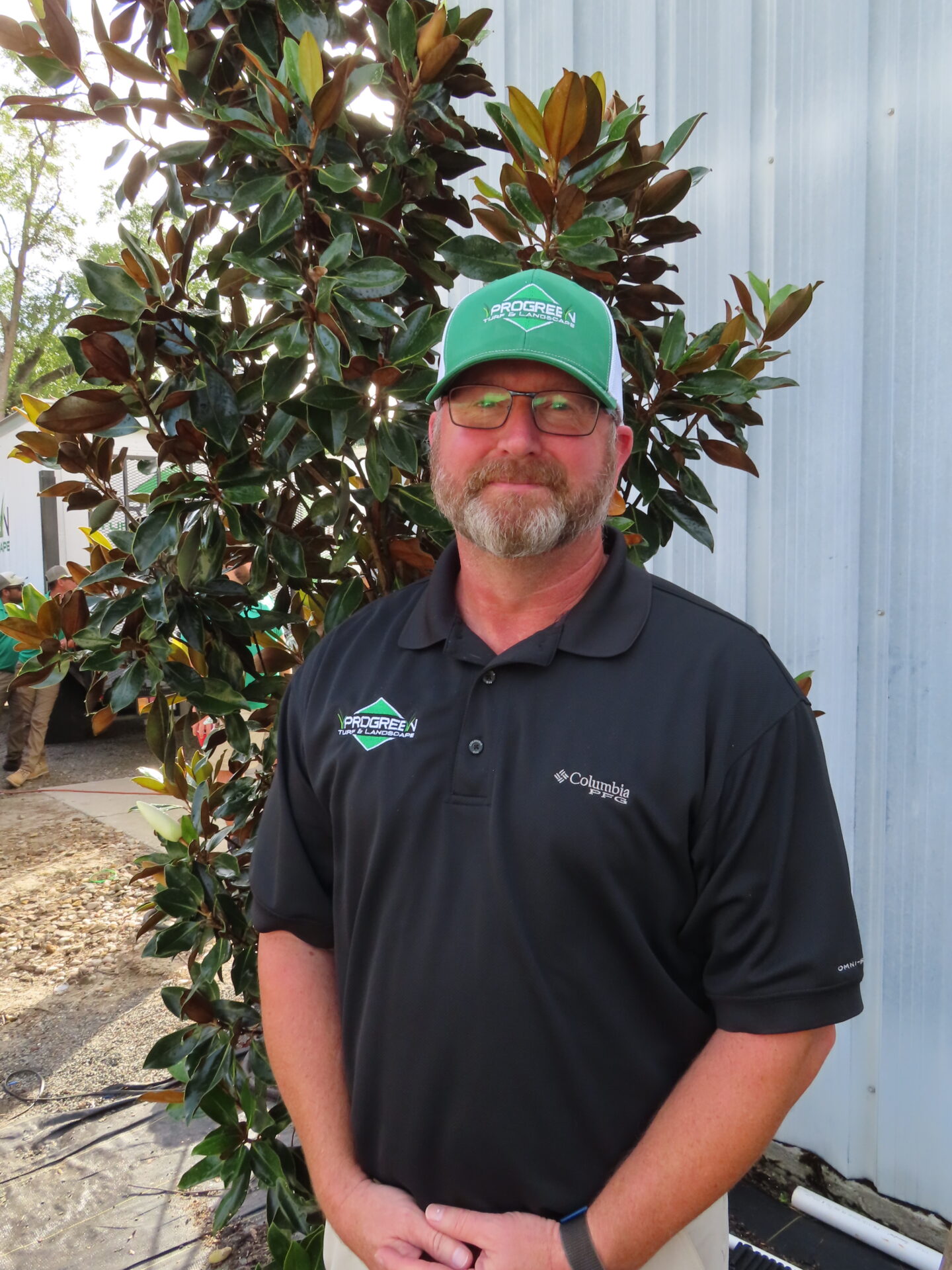 A person wearing a green cap with logo stands near a leafy plant, in front of a metal structure, smiling casually.